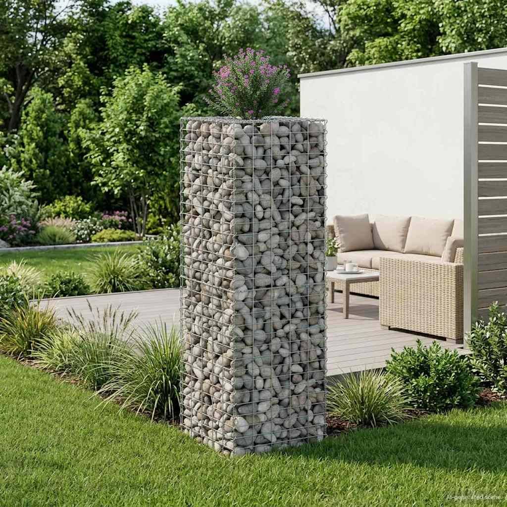 Ein Garten mit einer mit Steinen gefüllten Gabionensäule, die mit violetten Blumen gekrönt ist. Im Hintergrund befindet sich auf einer Terrasse ein Korbsofa-Set.