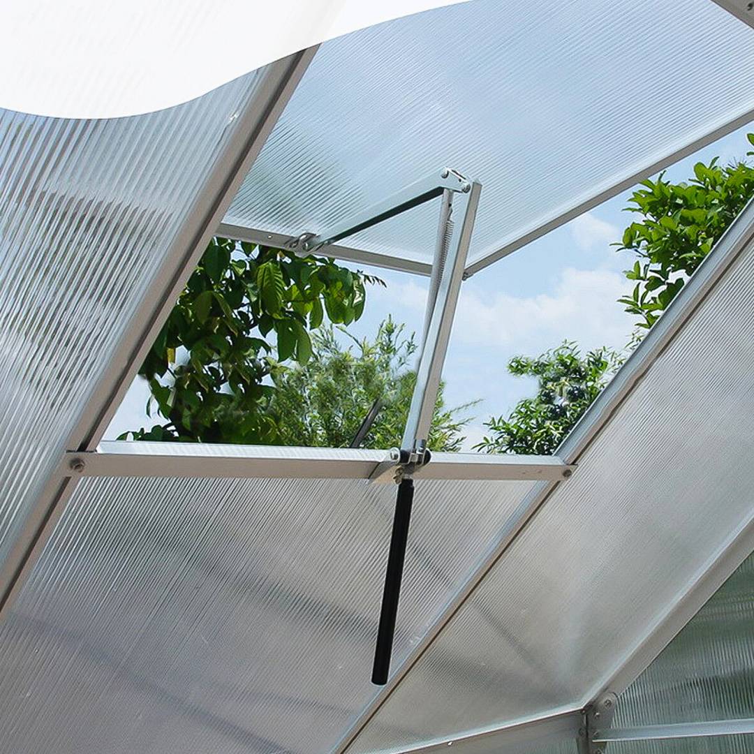 Offene Dachentlüftung in einem durchscheinenden Gewächshausdach, hochgestellt, um Luft und Licht hereinzulassen, mit Bäumen und blauem Himmel sichtbar außerhalb.