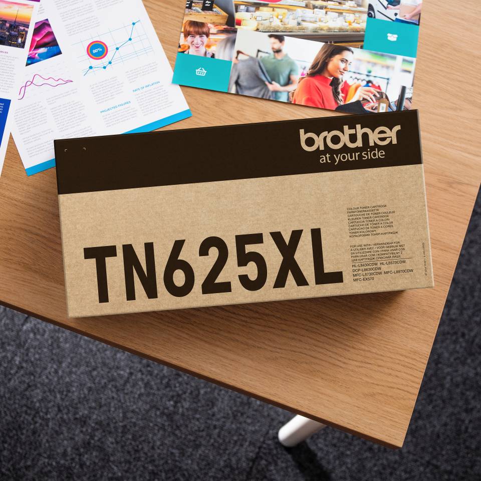 Brother TN625XL Druckertonerkartuschenbox auf einem Holzschreibtisch, mit Broschüren und Berichten im Hintergrund.