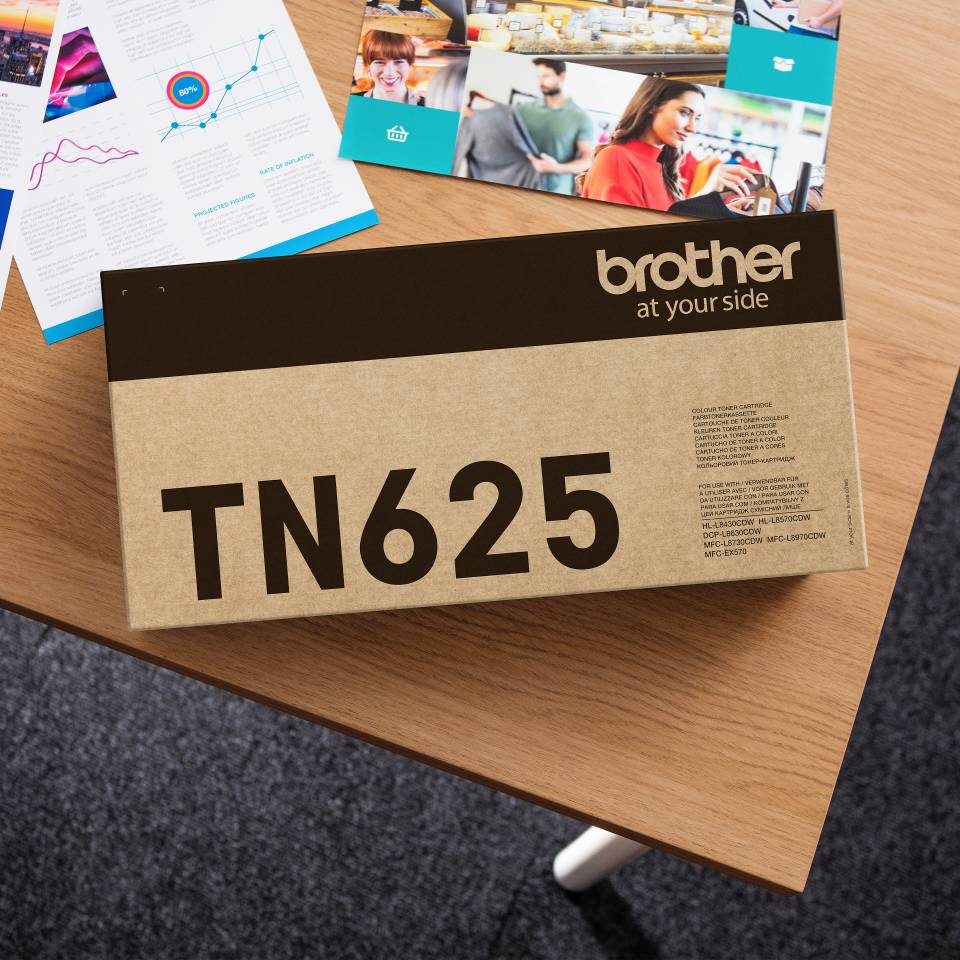 Brother TN625 Tonerpatrone in einer Schachtel auf einem Schreibtisch, mit Broschüren und gedruckten Berichten im Hintergrund.