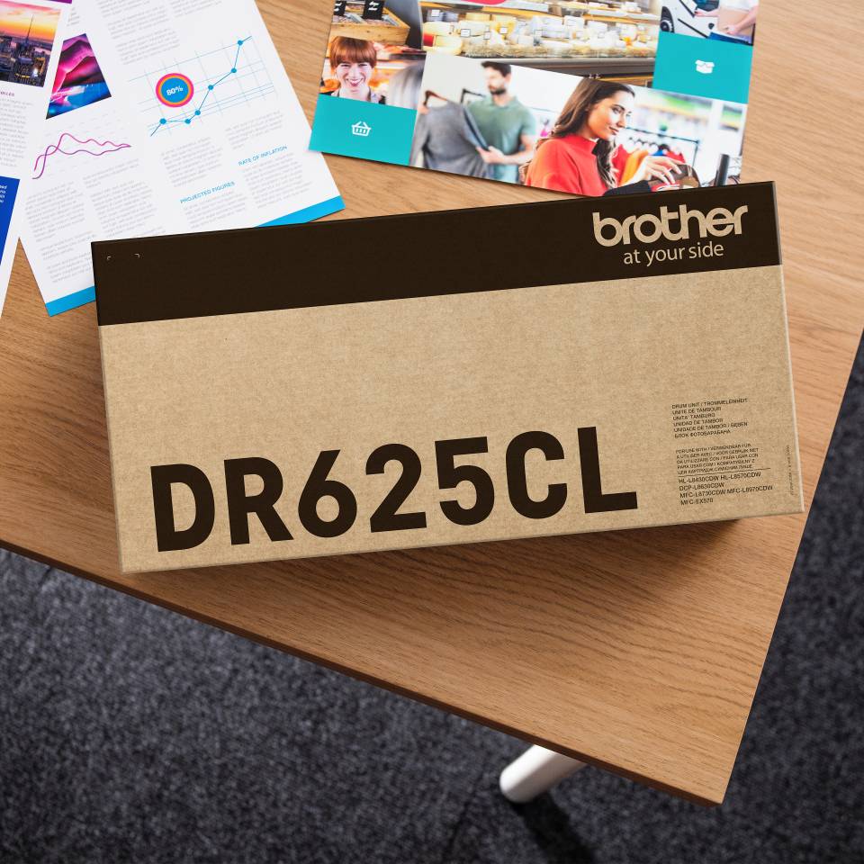 Tonerpatrone Brother DR625CL in einer Schachtel auf einem Holztisch, mit Broschüren und Fotos im Hintergrund.