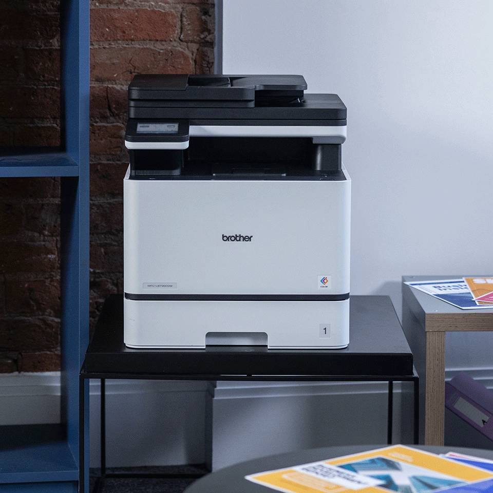 Brother Multifunktionsdrucker auf einem schwarzen Tisch in einem büroähnlichen Raum, mit Papieren in der Nähe und Ziegeln-Weiß-Wänden im Hintergrund.