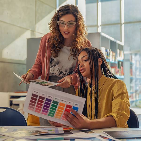 Zwei Frauen arbeiten zusammen an einem Tisch und überprüfen gedruckte Farbmuster und Designmuster in einem hellen Studio oder Klassenzimmer.