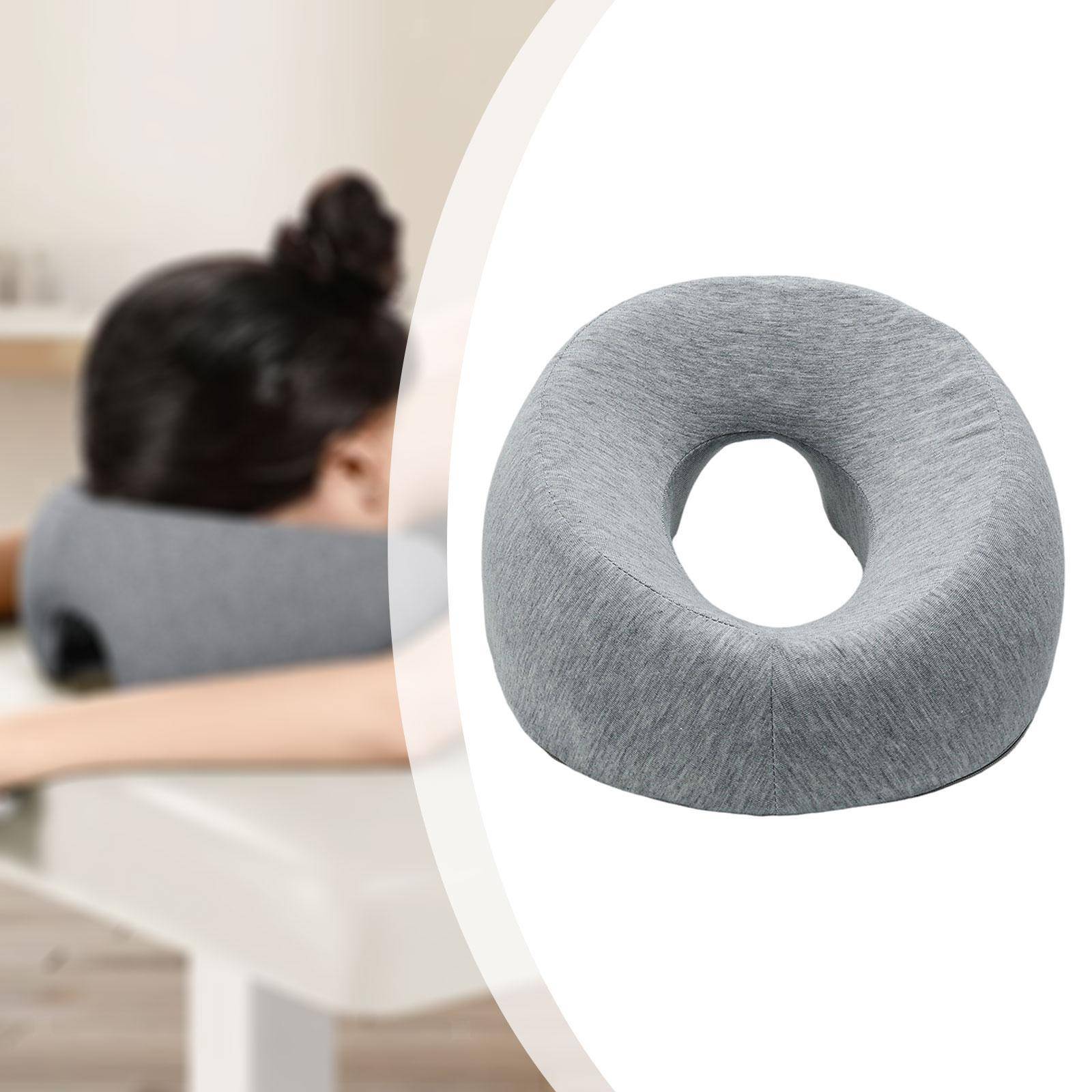 Grau geformtes Nackenkissen mit Donutform im Vordergrund, mit einer Person, die auf einem Bett im Hintergrund ruht.