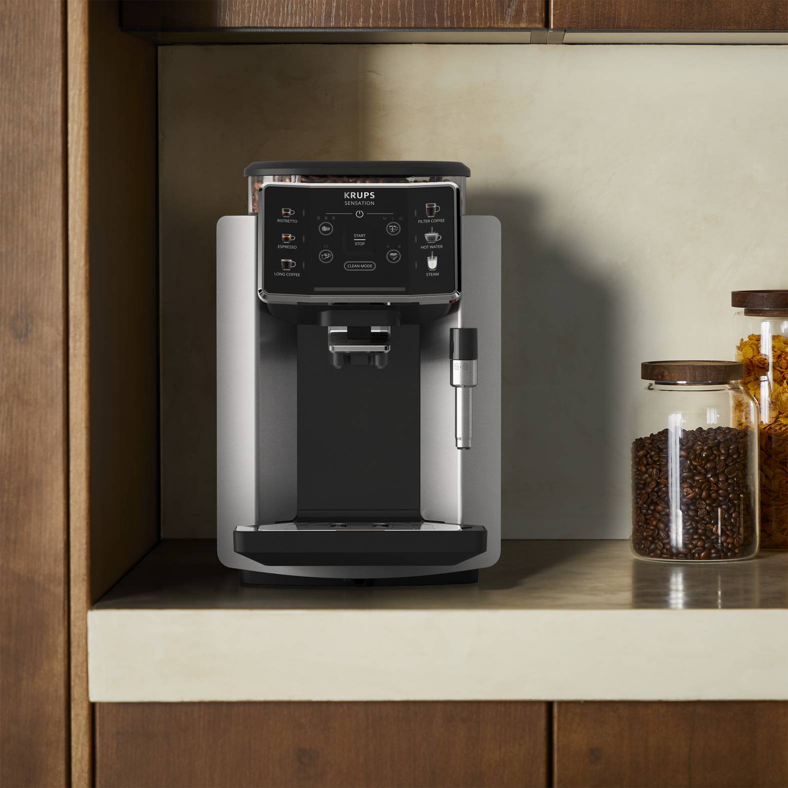 Silberne und schwarze KRUPS-Kaffeemaschine auf einem Küchenbord neben Gläsern mit Kaffeebohnen und Zuckerwürfeln.