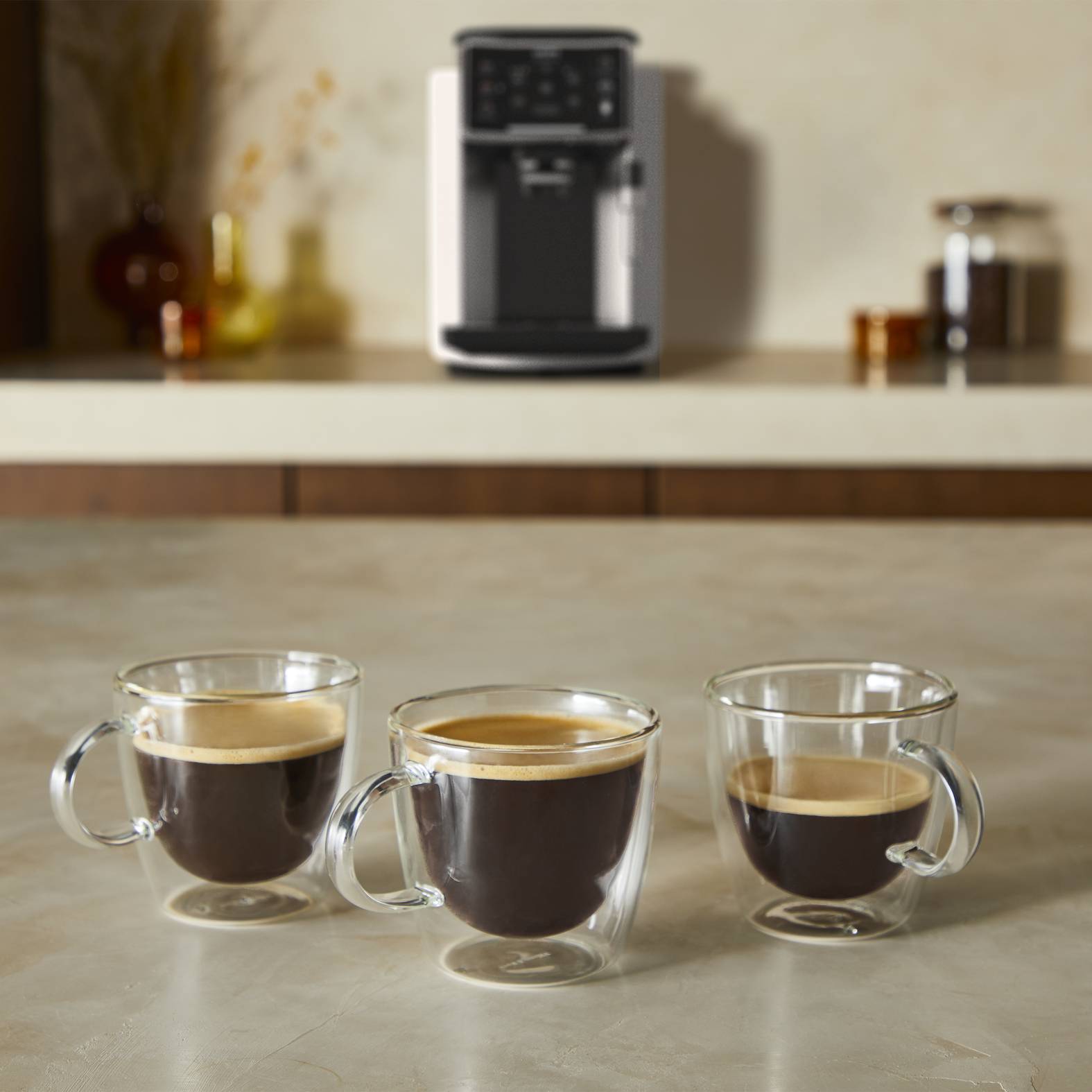 Drei doppelwandige Glastassen mit Espresso stehen auf einer Arbeitsplatte, wobei eine Kaffeemaschine verschwommen im Hintergrund zu sehen ist.