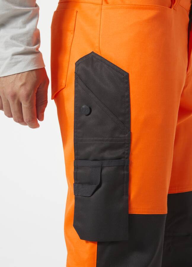 Nahaufnahme einer orangefarbenen Arbeitshose mit einer schwarzen Cargotasche am Oberschenkel, die Details der Nutzungstasche und eine Hand daneben zeigt.