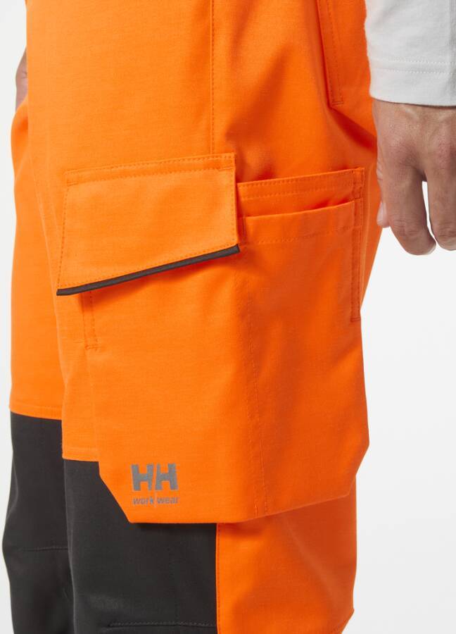 Nahaufnahme einer orangefarbenen Arbeitshose mit einer großen Cargotasche und schwarzem Besatz, getragen von einer Person, die vor einem weißen Hintergrund steht.