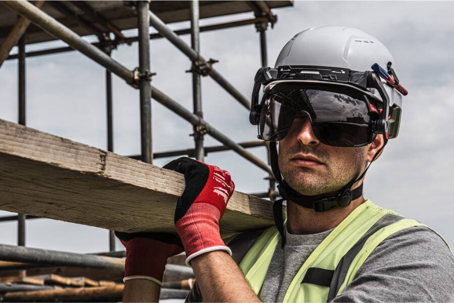 Bauarbeiter in Schutzhelm und Schutzbrille trägt ein Holzbrett auf einer Gerüstbaustelle.