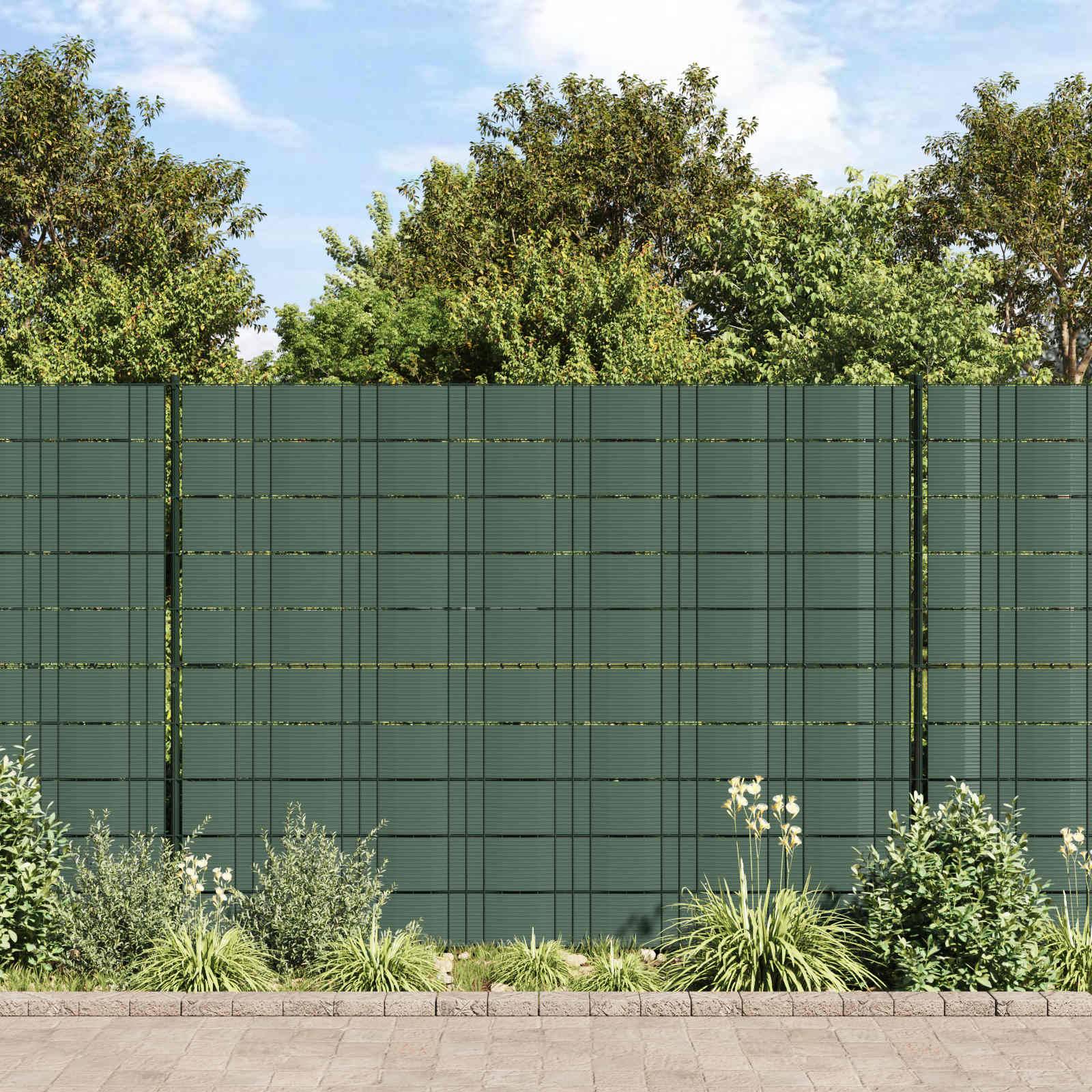 Hoher grüner Sichtschutzzaun hinter einem gestalteten Gartenbeet mit Sträuchern und Gräsern, mit Bäumen und blauem Himmel darüber.
