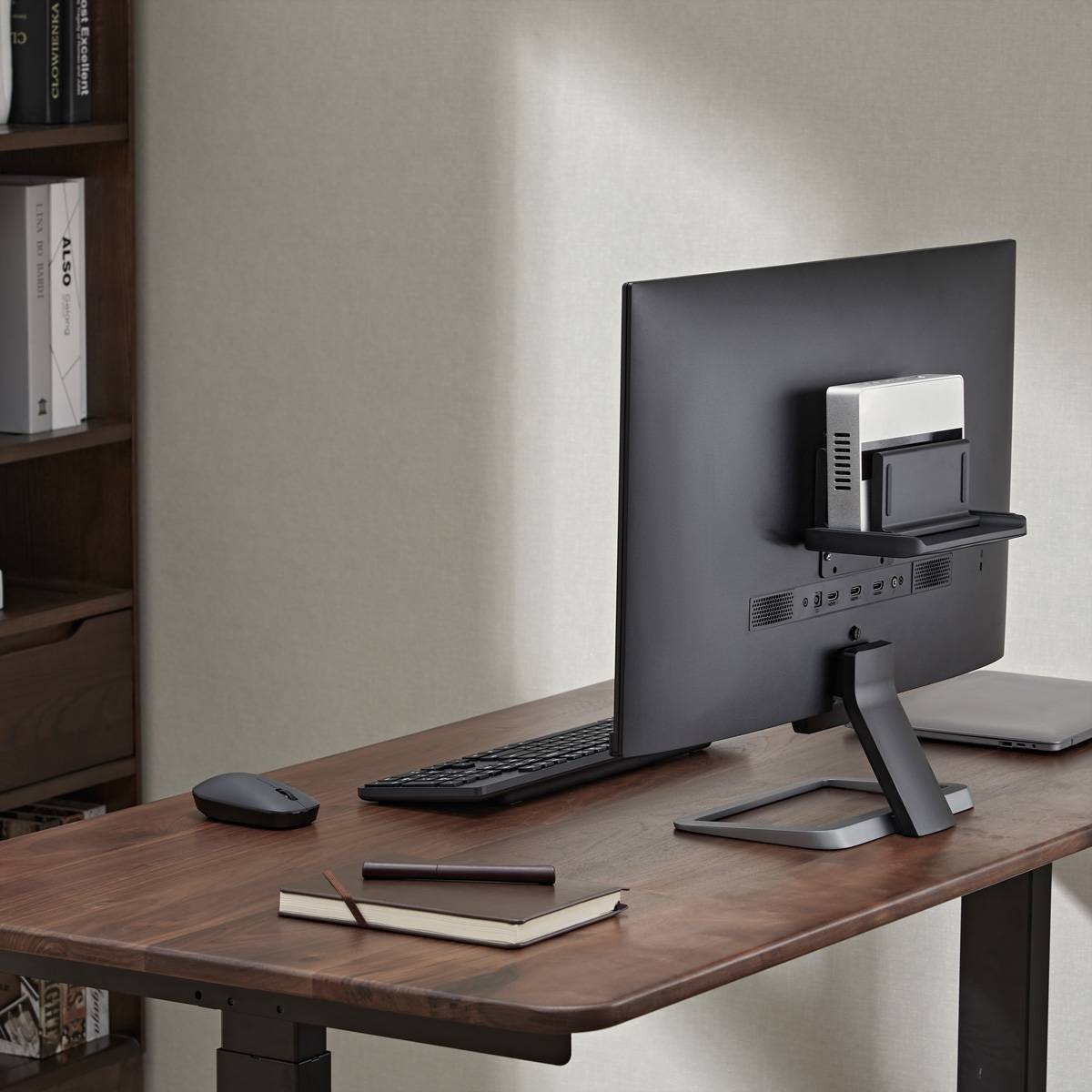 Arbeitszimmer-Schreibtisch mit einem Desktop-Monitor, Tastatur, Maus, Notizbuch und Stift neben einem Bücherregal.