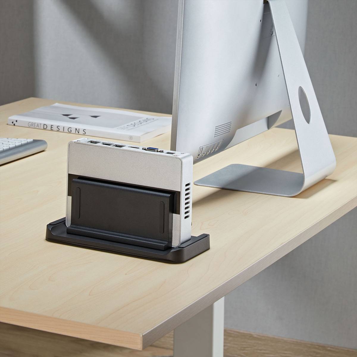 Ein kleiner Desktop-Ständer hält ein Gerät oder Tablet aufrecht auf einem Holzschreibtisch neben einem Computermonitor.