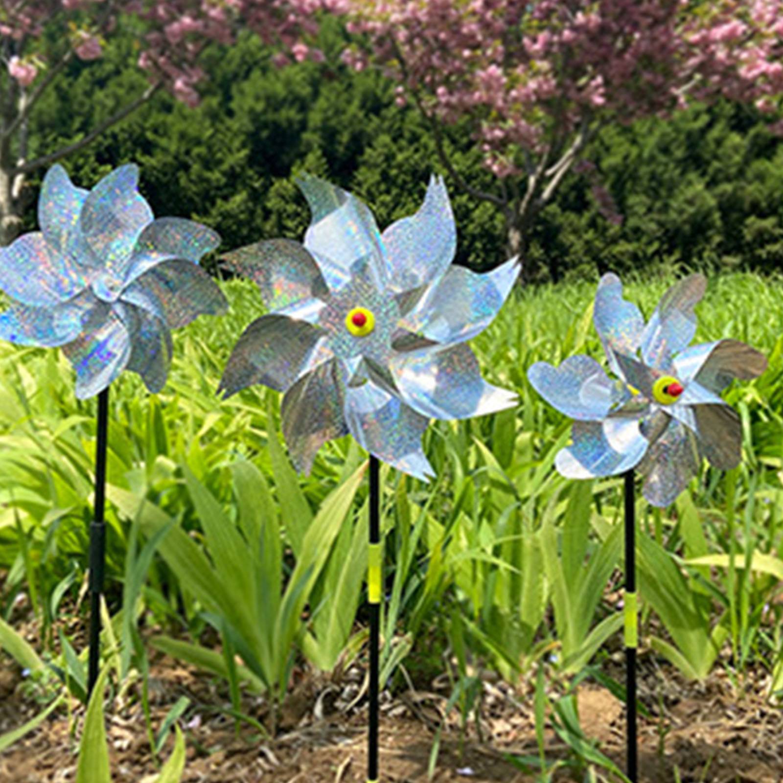 Drei silberne, windmühlenähnliche Gartenschmuckelemente auf Stangen stehen zwischen grünen Pflanzen in einem Blumenbeet, mit Bäumen und rosa Blüten im Hintergrund.