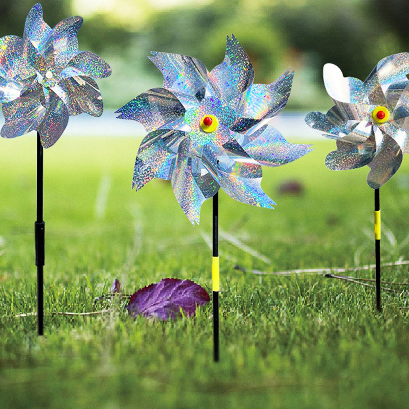 Drei schillernde Windmühlen-Gartenstecker drehen sich in einem grasbewachsenen Hof, mit einem violetten Blatt im Vordergrund.