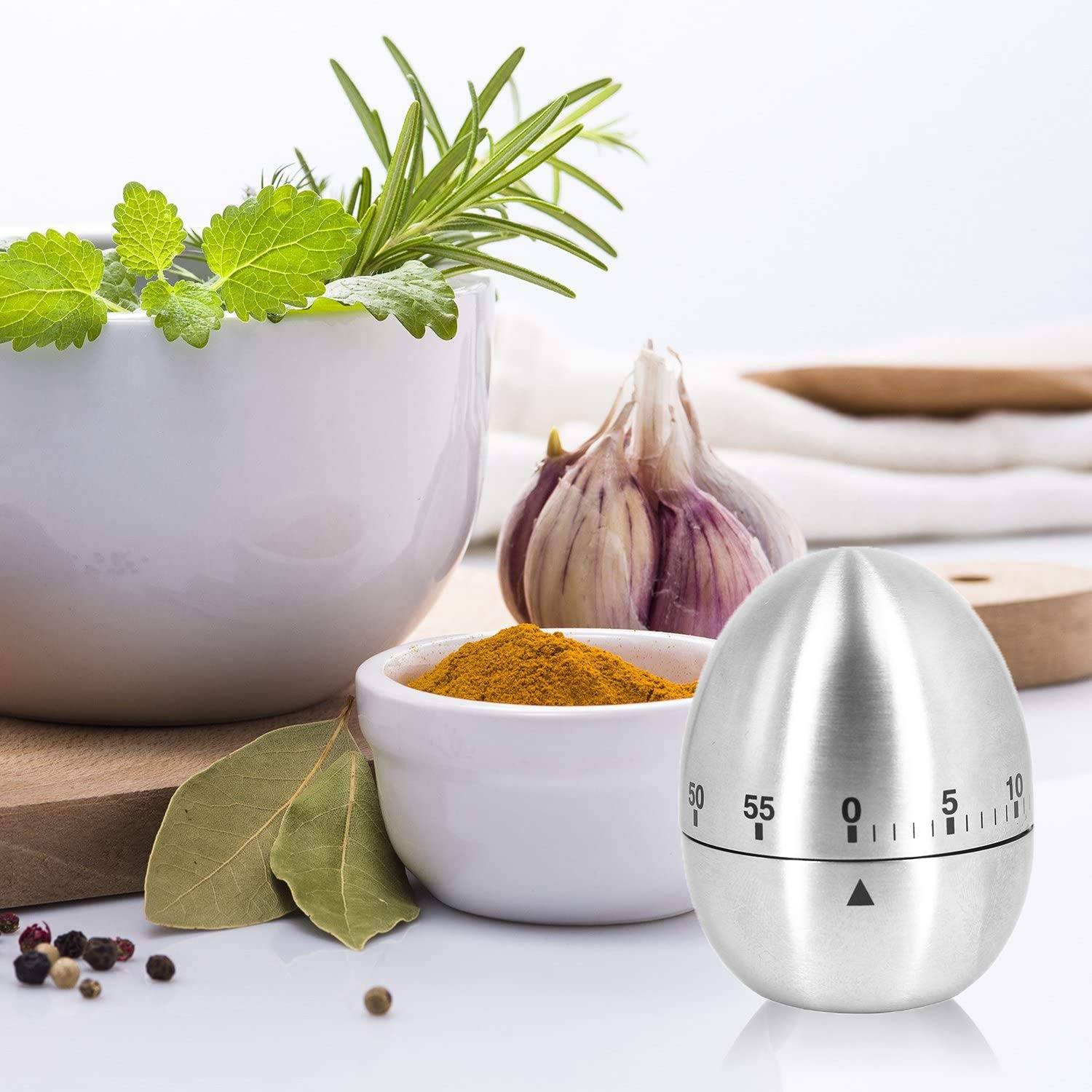 Stillleben in der Küche mit Kräutern, Knoblauch, Gewürzen, Lorbeerblättern und einem metallenen eiförmigen Timer im Vordergrund.