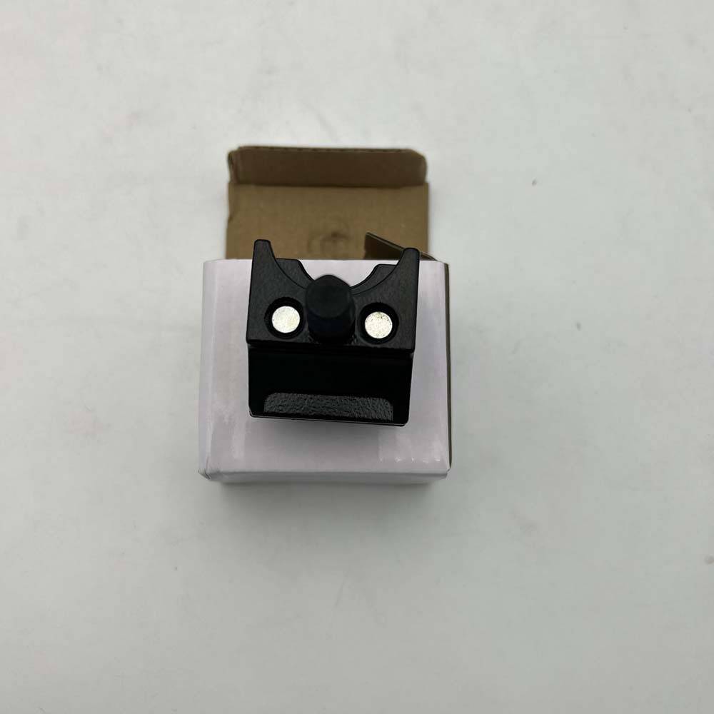 Schwarze Kunststoff-Montageklammer mit zwei silbernen Befestigungselementen sitzt auf einer kleinen weißen Schachtel vor einfachem Hintergrund.