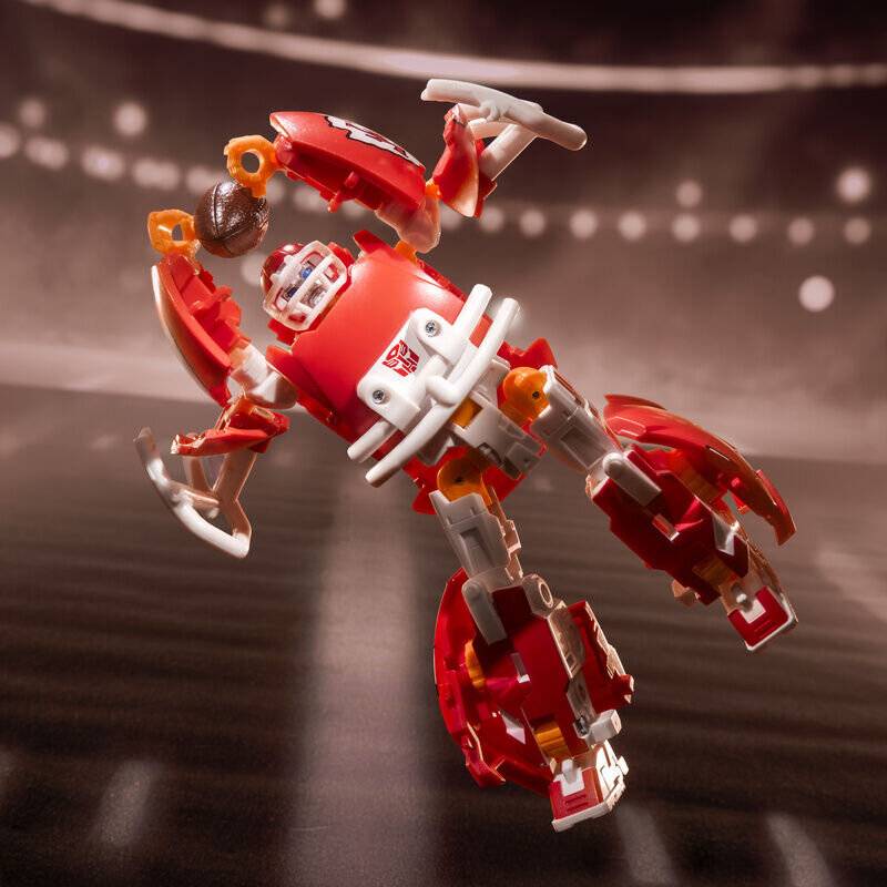 Rotes und weißes Roboter-Spielzeug, in der Luft schwebend, mit klauenartig geformten Gliedmaßen und kompaktem Körper, vor einem verschwommenen Stadion-ähnlichen Hintergrund.