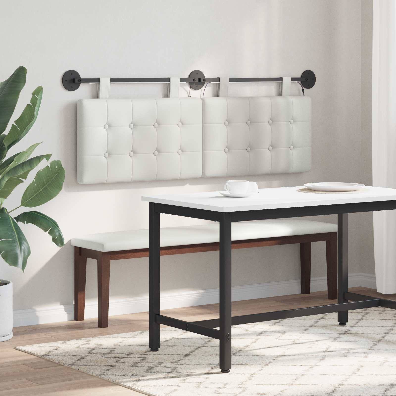 Modernes Esszimmer mit einer weißen gesteppten Wandpolsterung und Bank hinter einem Tisch mit schwarzem Rahmen auf einem hellen Teppich, neben einer Topfpflanze.