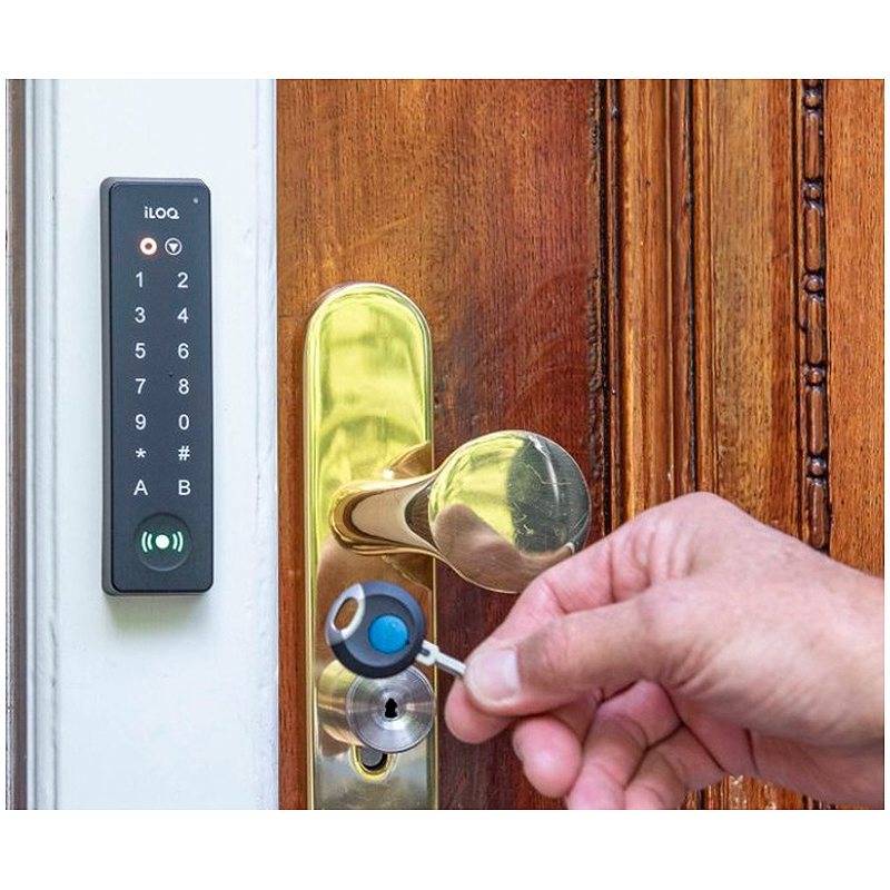 Eine Hand, die einen Schlüssel in ein Messingschloss an einer Tür einführt, neben einem wandmontierten iLOQ-Keypad-Zugangsfeld an einer Holztür.
