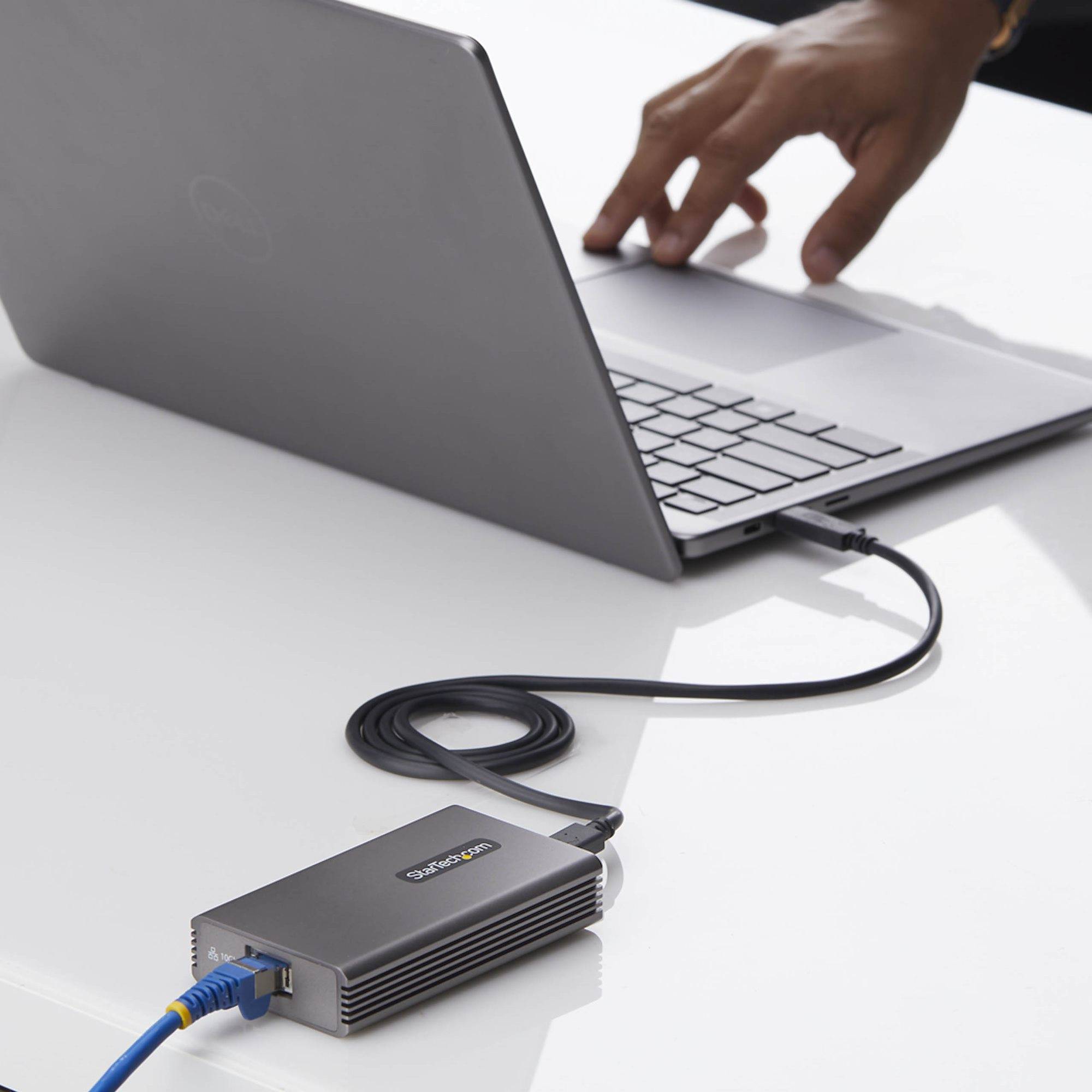 Laptop, das über ein Kabel mit einem kleinen externen Adapter auf einem Schreibtisch verbunden ist, mit einer Hand, die das Laptop-Trackpad benutzt.