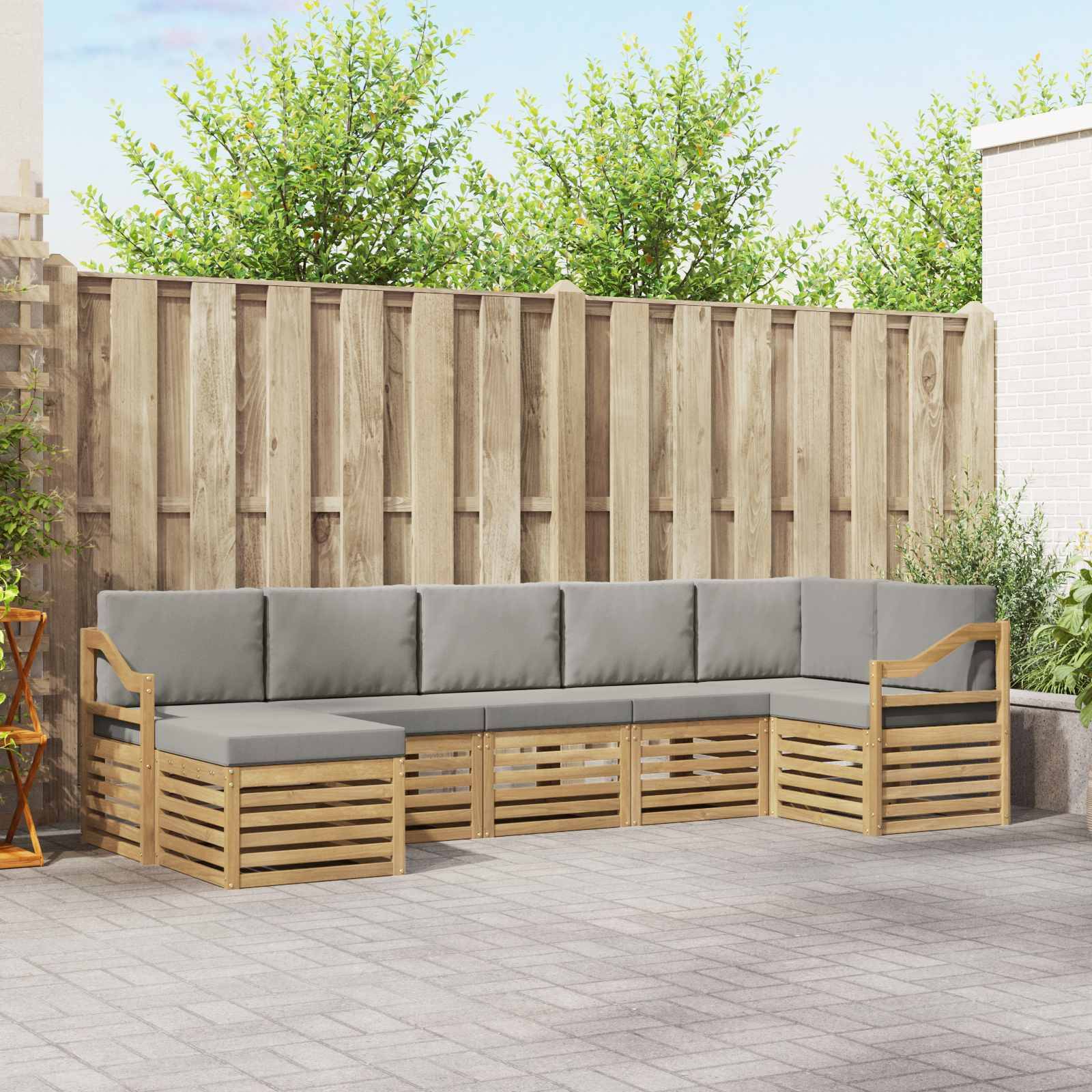 Outdoor L-förmiges Holz-Ecksofa mit grauen Kissen auf einer Terrasse, vor einem hohen Zaun und Grünflächen.