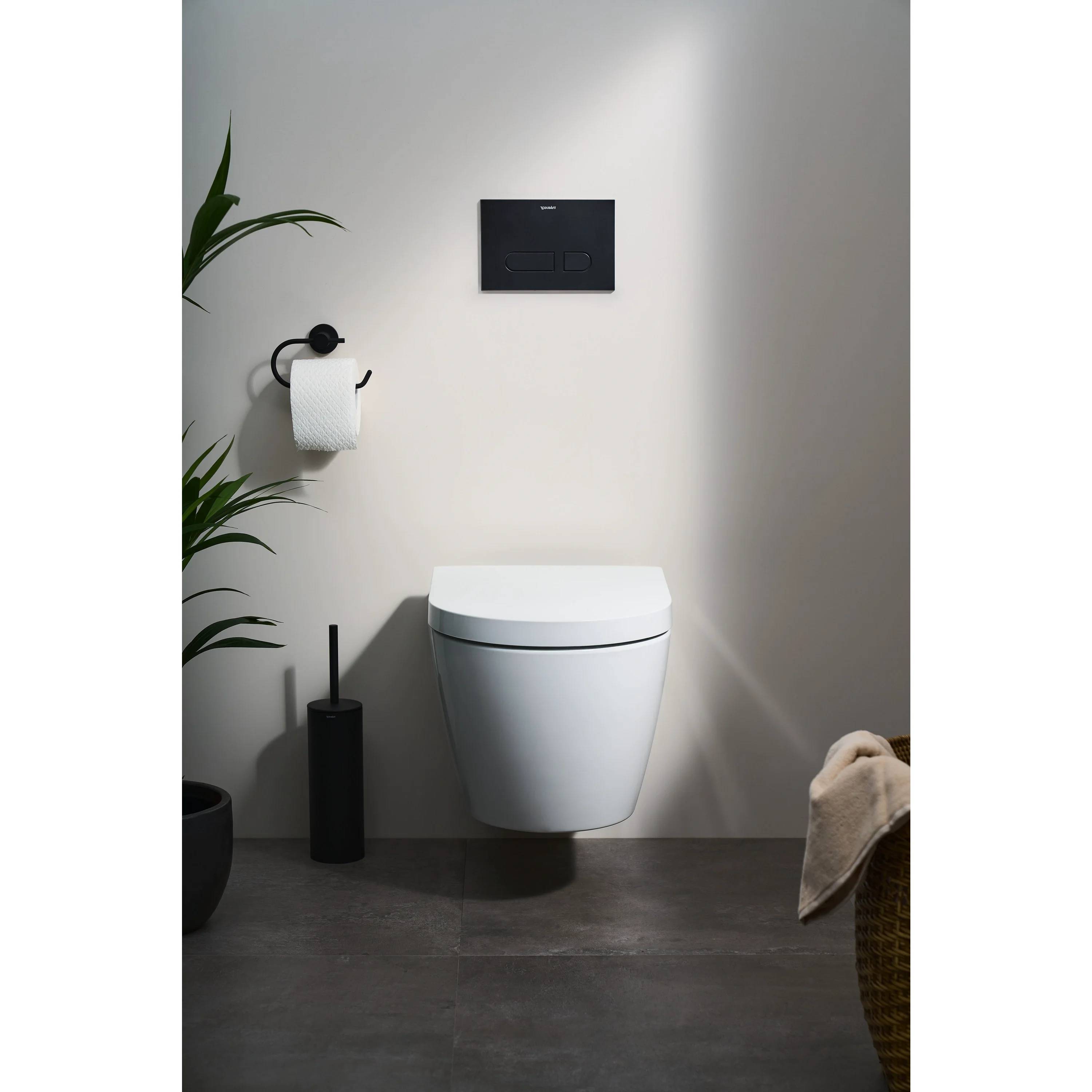Modernes Badezimmer mit einer wandmontierten weißen Toilette, schwarzem Spülpanel, Toilettenpapierhalter, Pflanze und Wäschekorb auf grauem Fliesenboden.