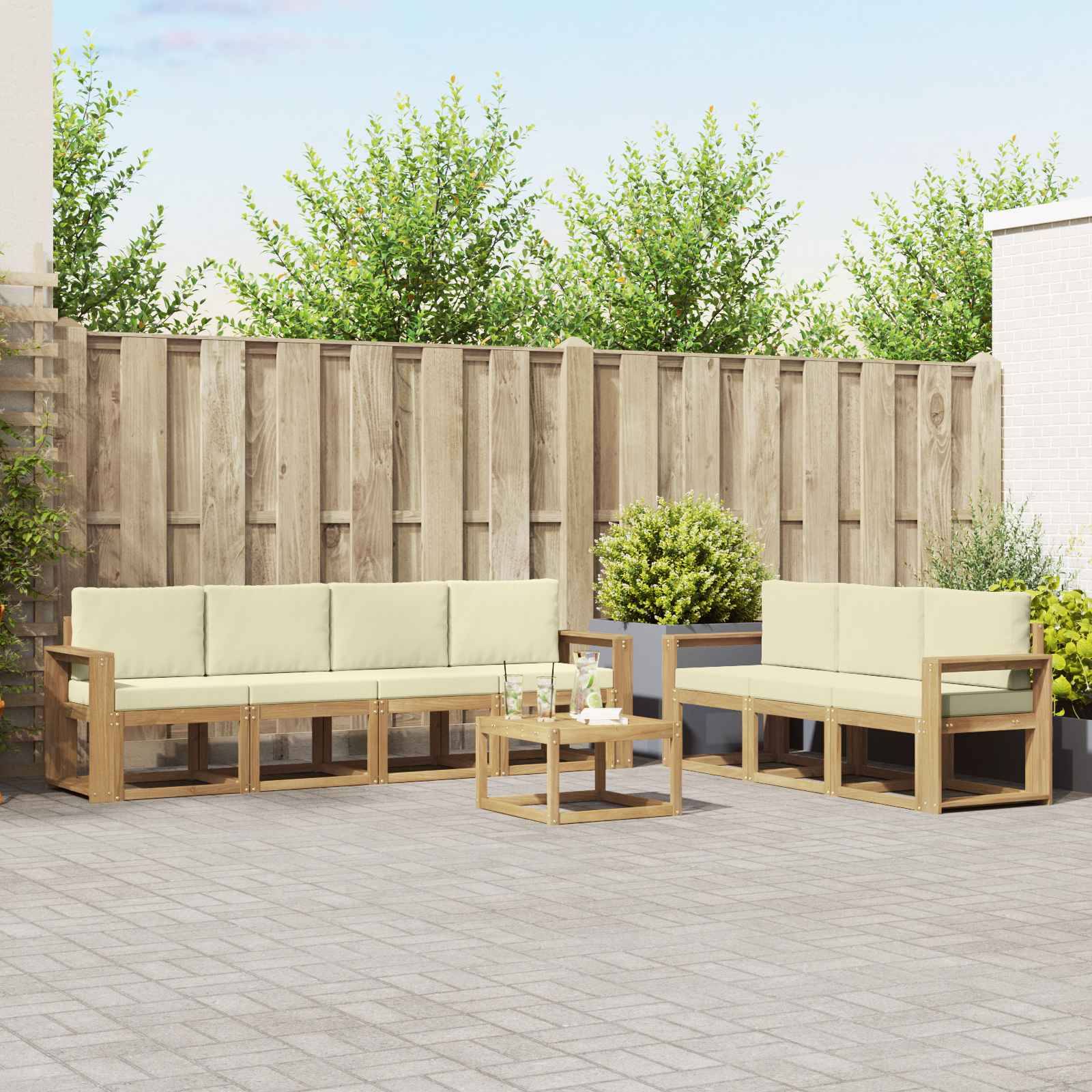 Außenpatio mit zwei hellem Holzsofa und einem kleinen Glastisch auf einer gepflasterten Terrasse, vor einem Holzzaun und Sträuchern.