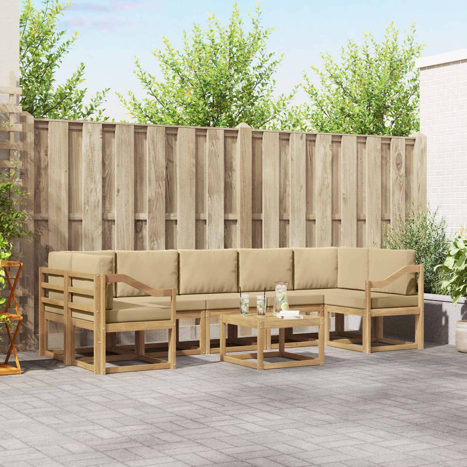 Outdoor-Sitzgruppe auf der Terrasse mit beigen gepolsterten Holzlounge-Sofas und Couchtisch auf gepflasterter Terrasse, vor einem Holzzaun und Grünflächen.