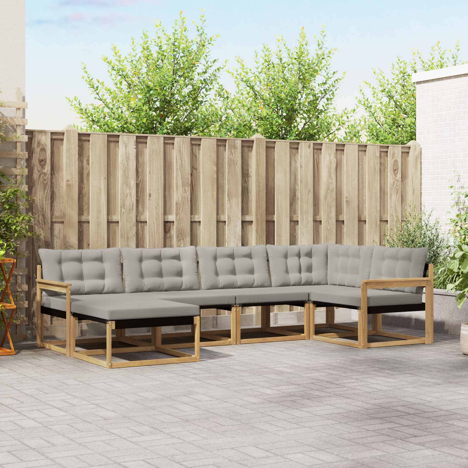 Graues modulares Outdoor-Ecksofa mit Holzrahmen und schwarzen Kissen auf einer gepflasterten Terrasse vor einem Holzzaun.