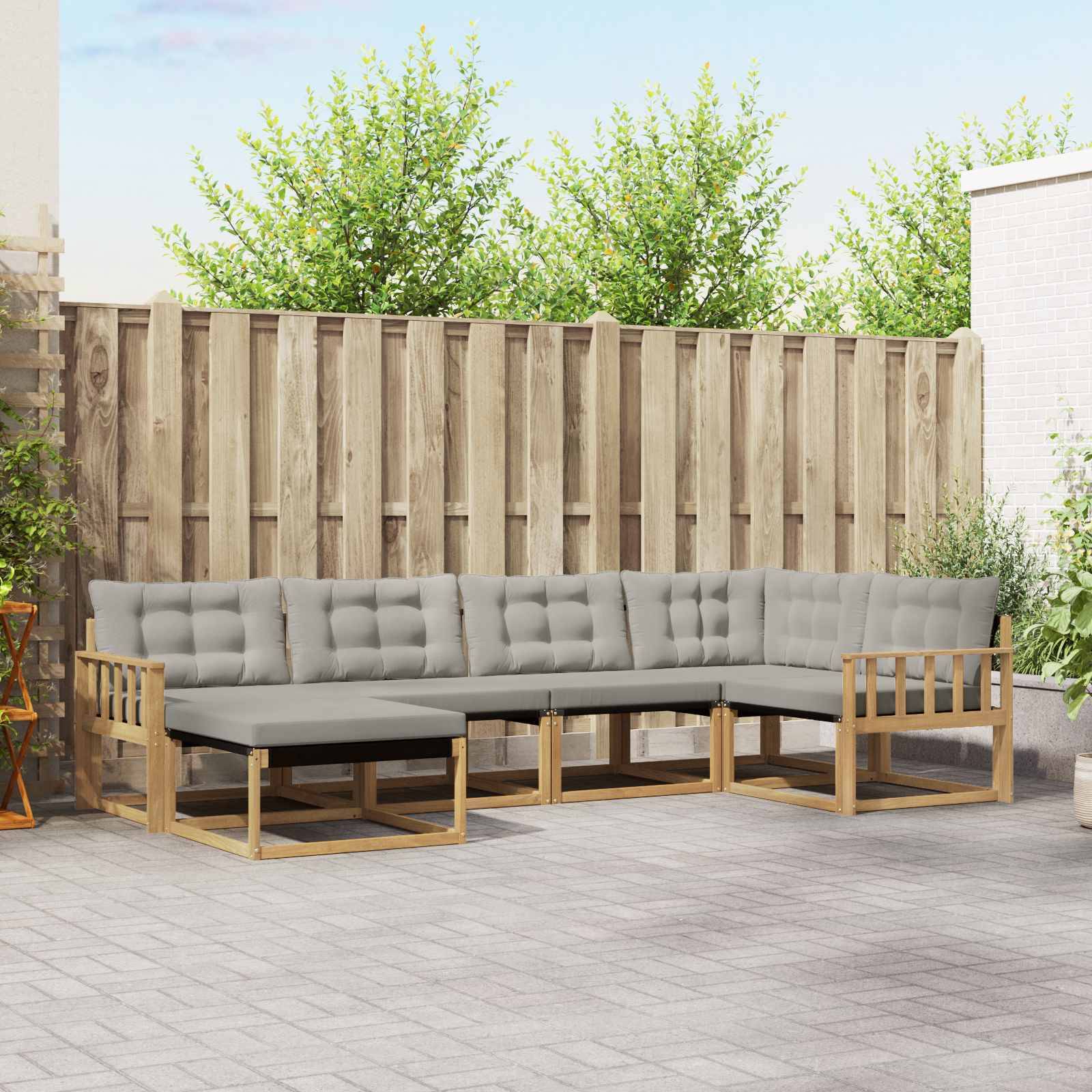 Outdoor-Terrassensektionalsofa mit grauen Kissen und Holzrahmen gegen einen Zaun in einem gepflasterten Hinterhof.