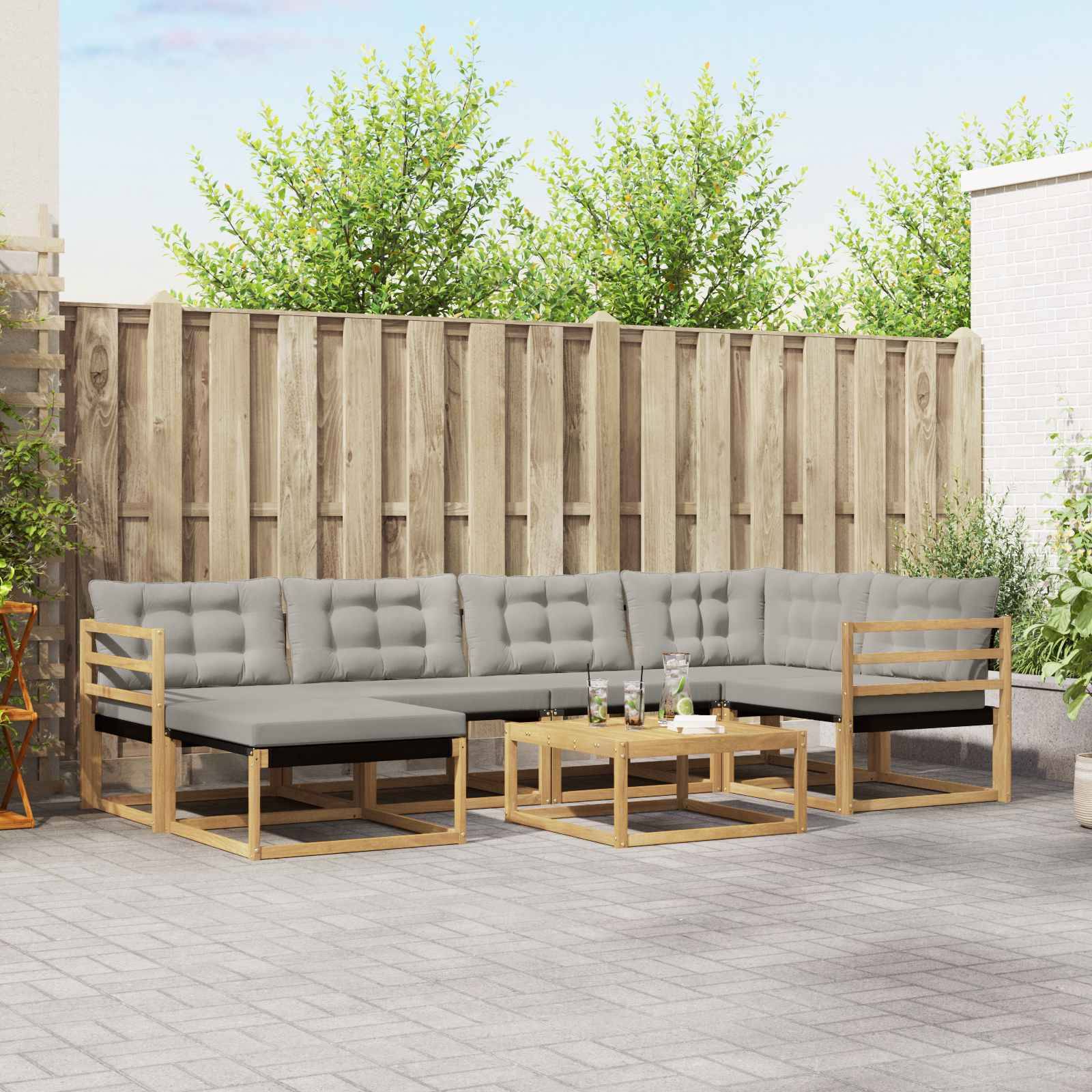 Outdoor-Terrasse mit grauem L-förmigem Ecksofa, passendem Hocker und Couchtisch vor einem Holzzaun.