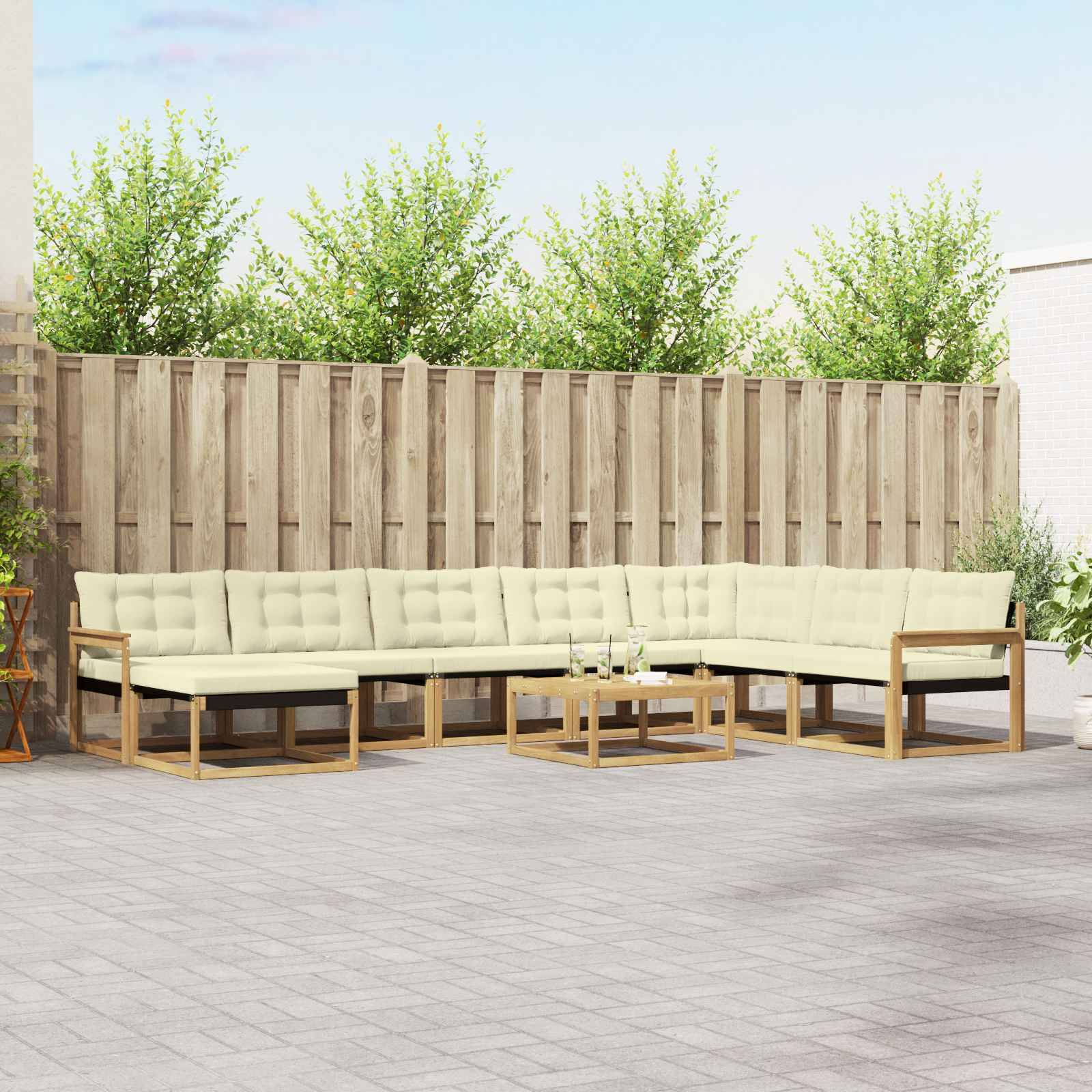 Outdoor-Terrassenbereich mit einem cremefarbenen Ecksofa und Glascouchtischen auf einer gekachelten Terrasse vor einem Holzzaun und grüner Bepflanzung.