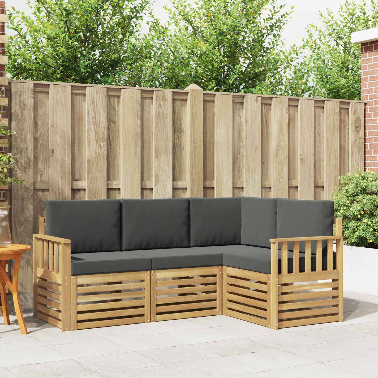Outdoor-Holz-L-förmiges Sitzkissen-Sofa mit dunklen Kissen auf einer Terrasse, vor einem hohen Holzzaun und Gartenbegrünung.