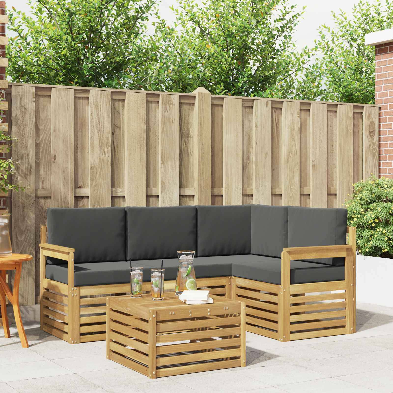 Outdoor-Terrasse mit einem schwarzgepolsterten Holz-Ecksofa und passendem Couchtisch, vor einem Holzzaun in einem Garten aufgestellt.