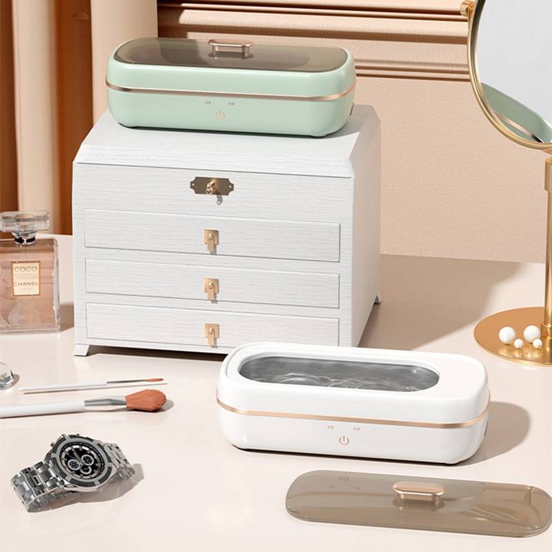 Eine Tischplattenausstellung von eleganten Beauty-Organizern und Geräten, einschließlich einer mintgrünen Schachtel auf einer weißen Schublade und einem weißen ovalen Gerät auf einem Waschtisch mit Make-up-Tools.
