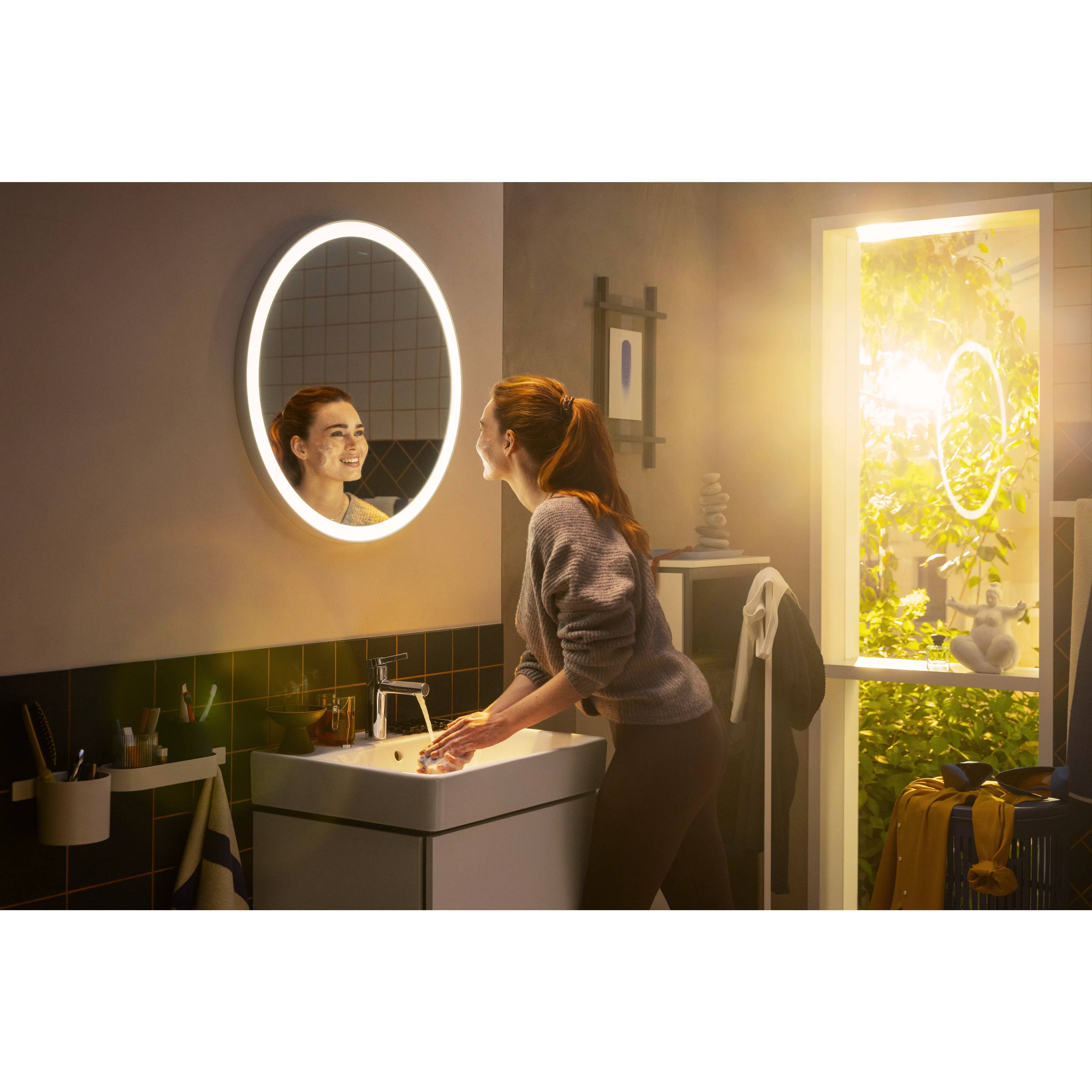 Eine Frau wäscht sich die Hände an einem Waschbecken im Badezimmer neben einem leuchtenden runden Spiegel, während Sonnenlicht durch eine offene Tür hereinströmt.