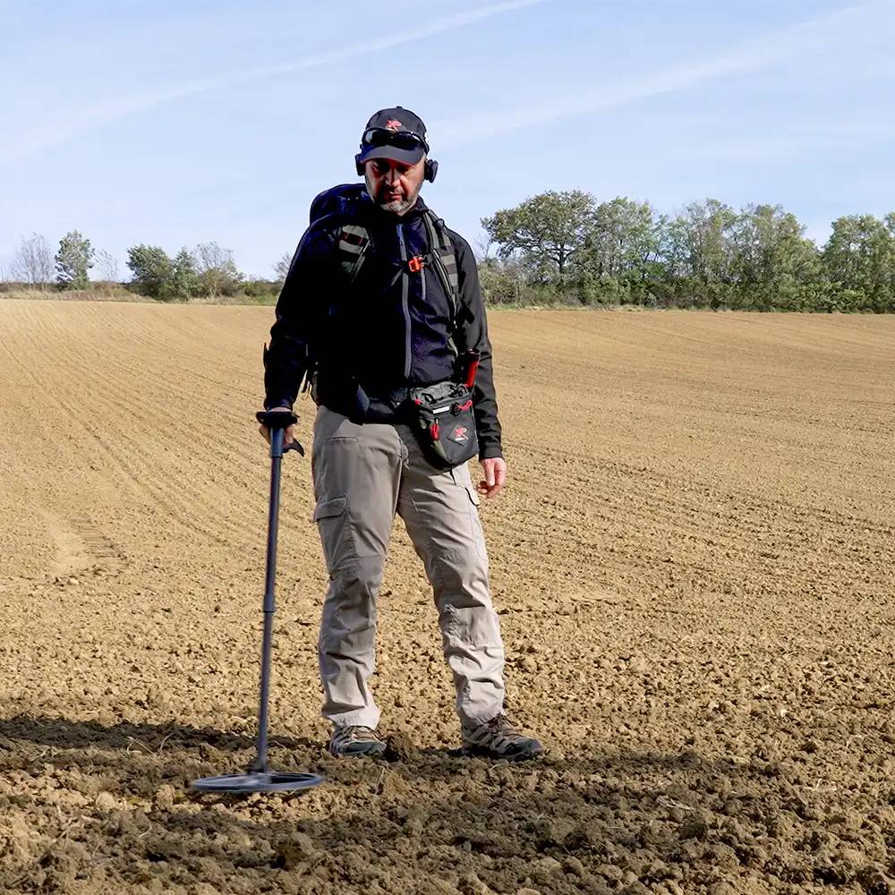 Ein Mann nutzt ein Metallsuchgerät auf einem gepflügten Feld an einem sonnigen Tag, trägt Outdoor-Ausrüstung und Kopfhörer.