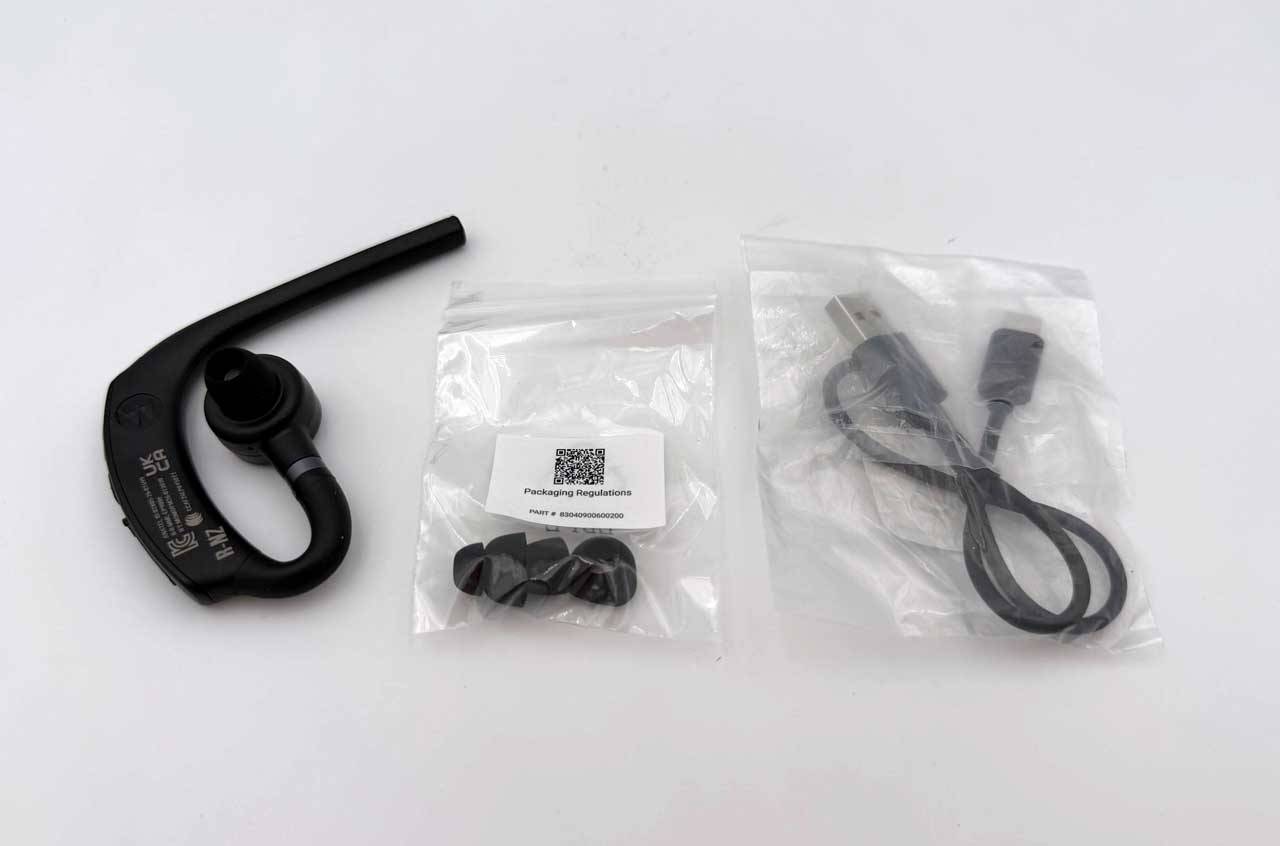 Schwarzes kabelloses Ohrhörer mit Ohrbügel neben zwei Plastiktüten mit Ohrstöpseln/Zubehör und einem USB-Ladekabel.