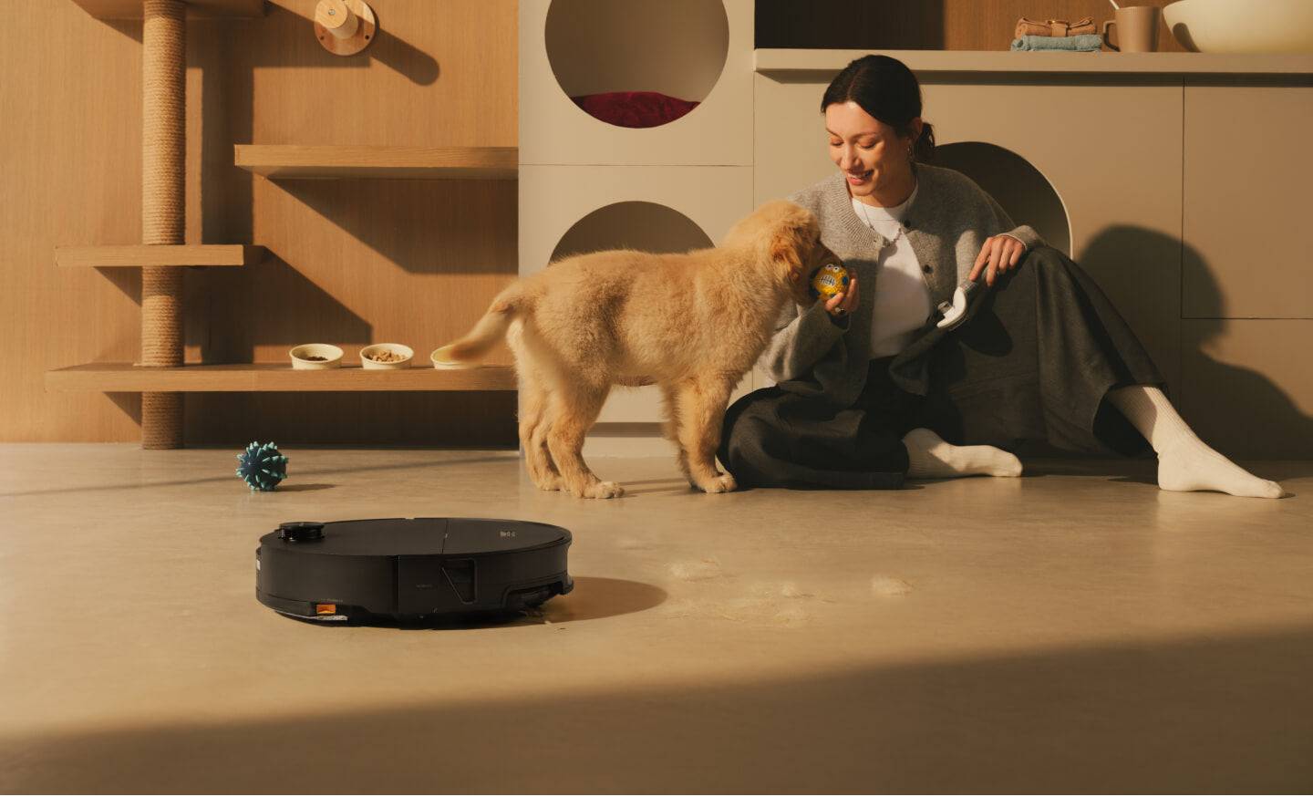 Eine Frau sitzt auf dem Boden und spielt mit einem Welpen, während ein Staubsaugerroboter in der Nähe in einem modernen Wohnzimmer reinigt.