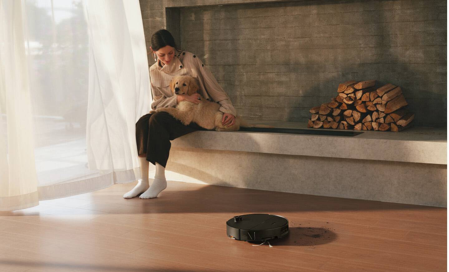 Eine Frau sitzt an einem Kamin und hält einen kleinen Hund, während ein Staubsaugerroboter vor ihr den Boden reinigt.