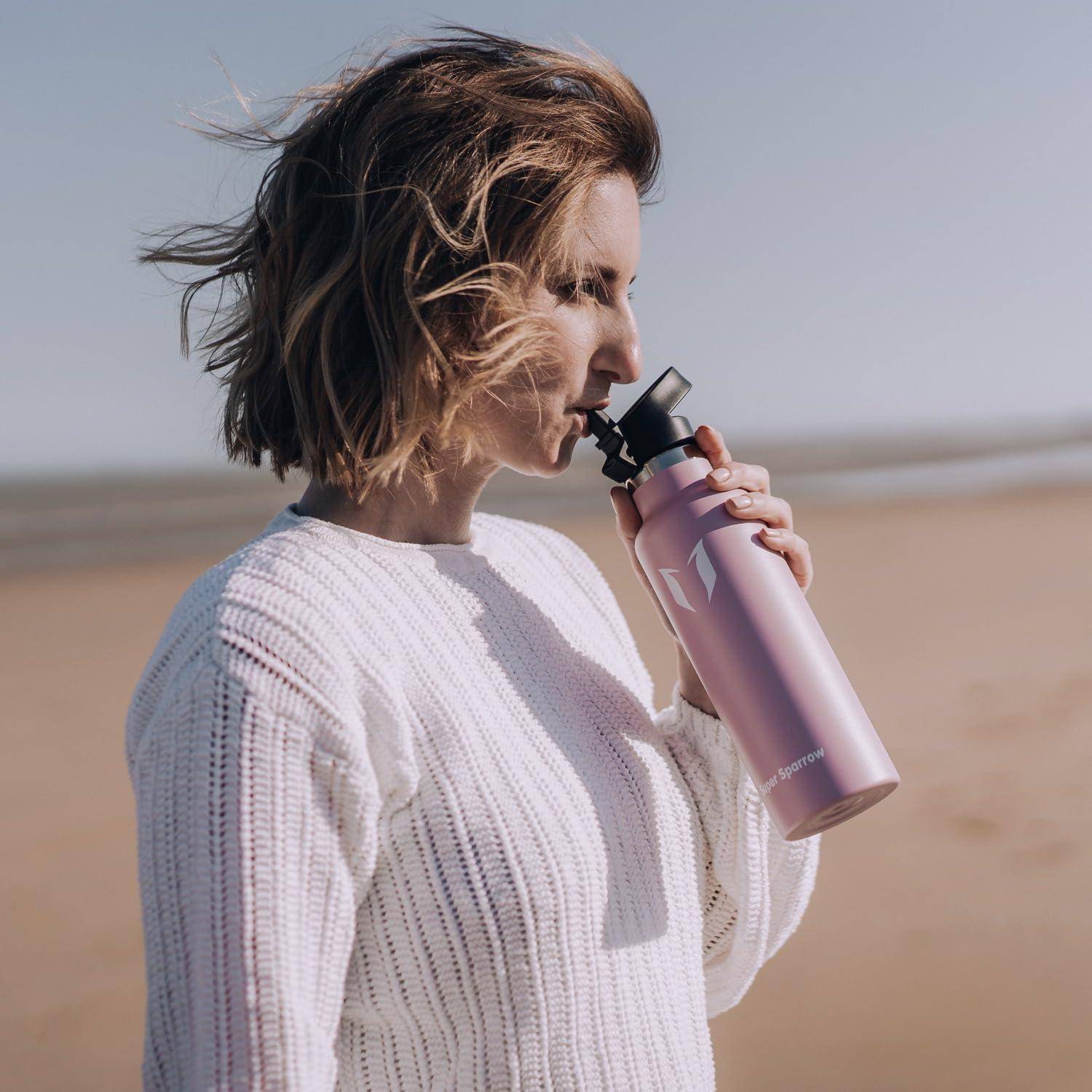 Frau in einem weißen Pullover trinkt aus einer rosa Wasserflasche am Strand, mit Sand und Ozean im Hintergrund.