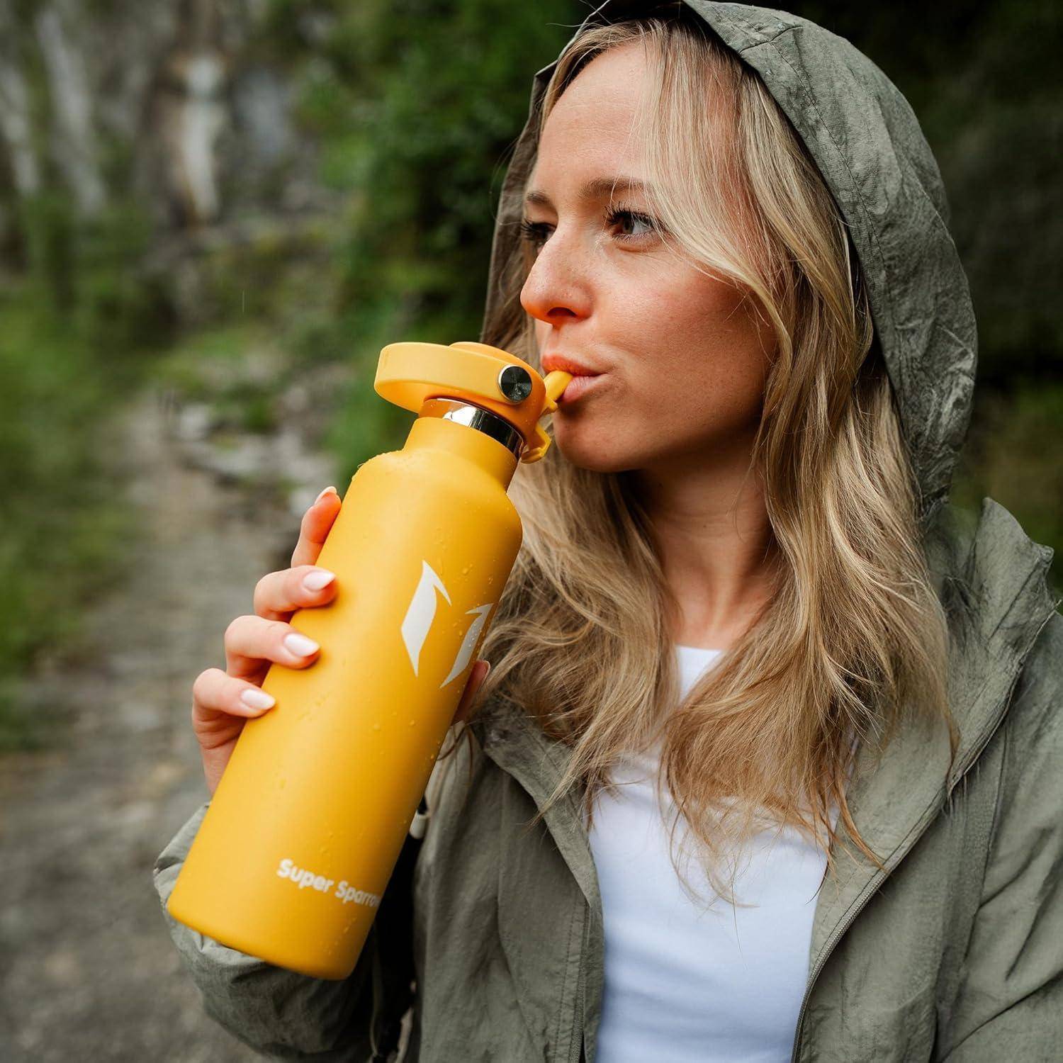 Frau in einer Jacke mit Kapuze trinkt aus einer leuchtend gelben Wasserflasche im Freien.