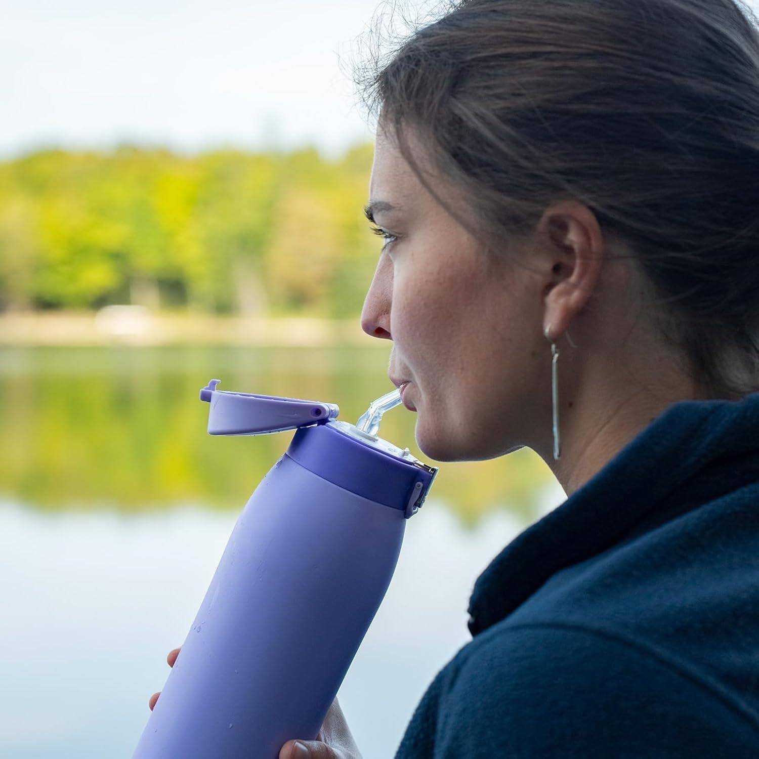 Frau trinkt aus einer violetten wiederverwendbaren Flasche am Seeufer, im Profil zu sehen, mit ruhigem Wasser und Bäumen im Hintergrund.