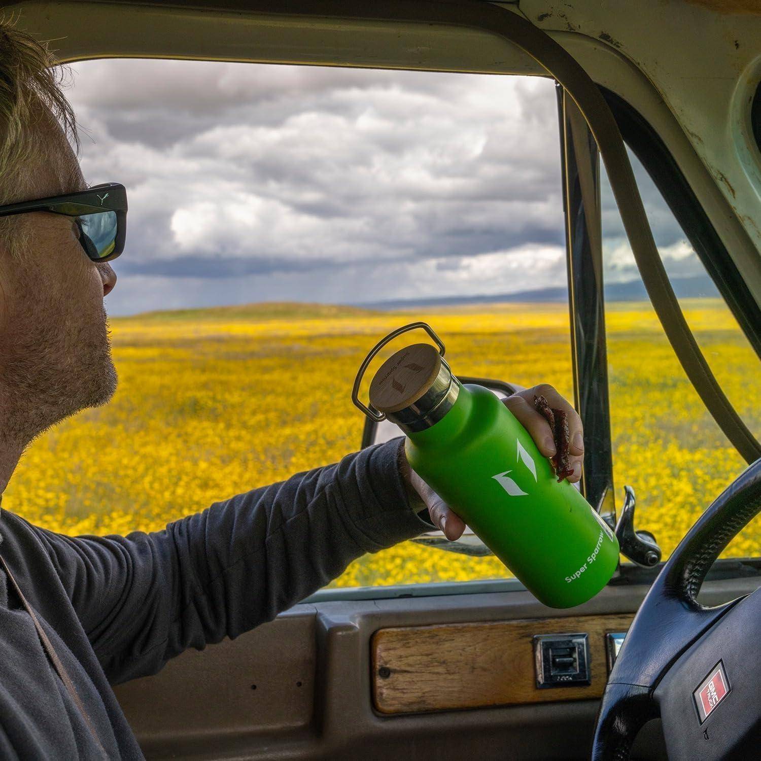 Ein Mann sitzt in einem Fahrzeug, hält eine grüne Wasserflasche und blickt auf ein gelbes Blumenfeld unter bewölktem Himmel.