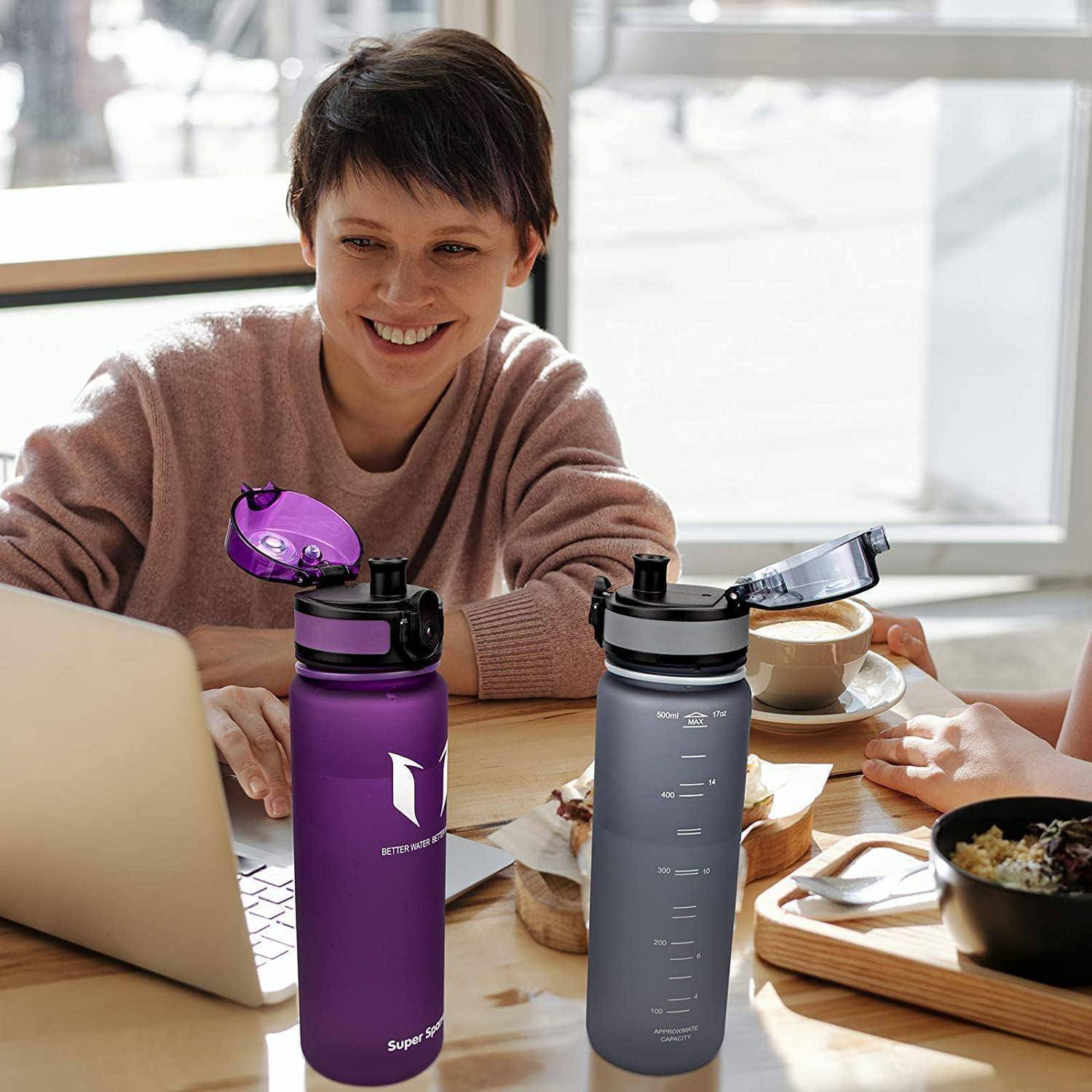 Lächelnde Frau an einem Tisch mit einem Laptop und Kaffee, die zwei wiederverwendbare Wasserflaschen in Lila und Grau mit Klappdeckeln zeigt.