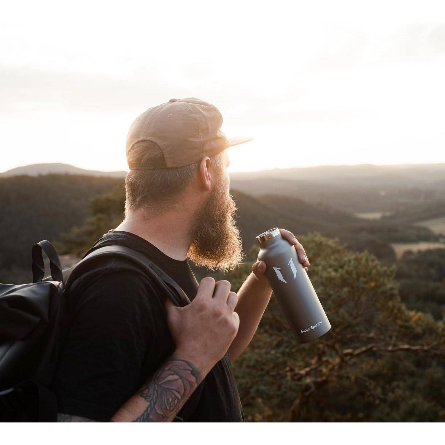 Bärtiger Mann mit Rucksack, der eine Wasserflasche hält und bei Sonnenuntergang auf eine hügelige Landschaft blickt.