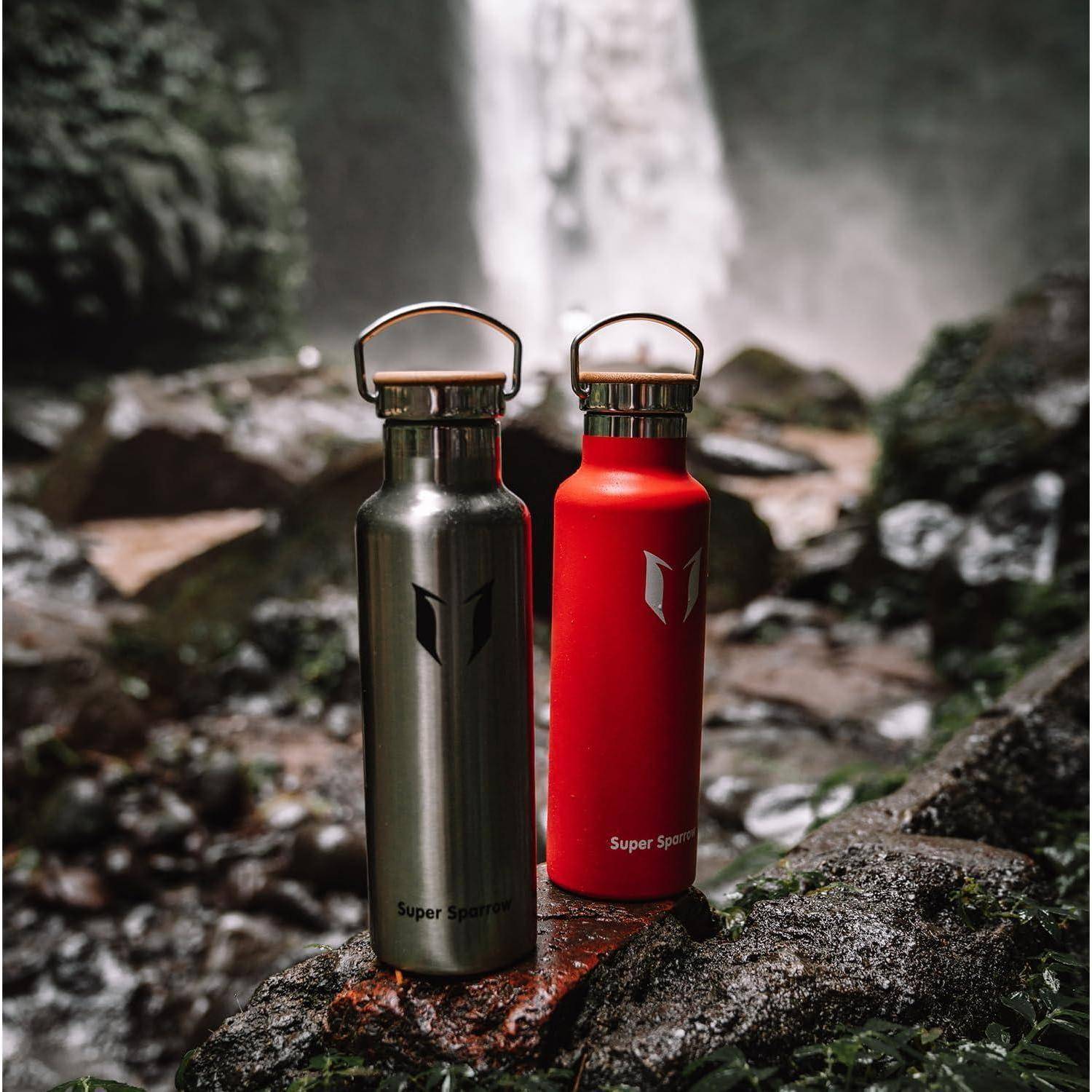 Zwei wiederverwendbare Metallwasserflaschen, eine schwarze und eine rote, stehen auf nassen Steinen in der Nähe eines Wasserfalls in einer Waldumgebung.