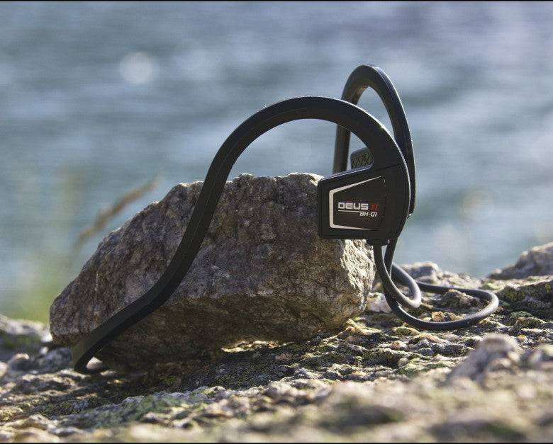 Schwarze wasserdichte Kopfhörer, die auf einem Stein am Wasser ruhen, mit dem Kabel, das um den Stein geschlungen ist.