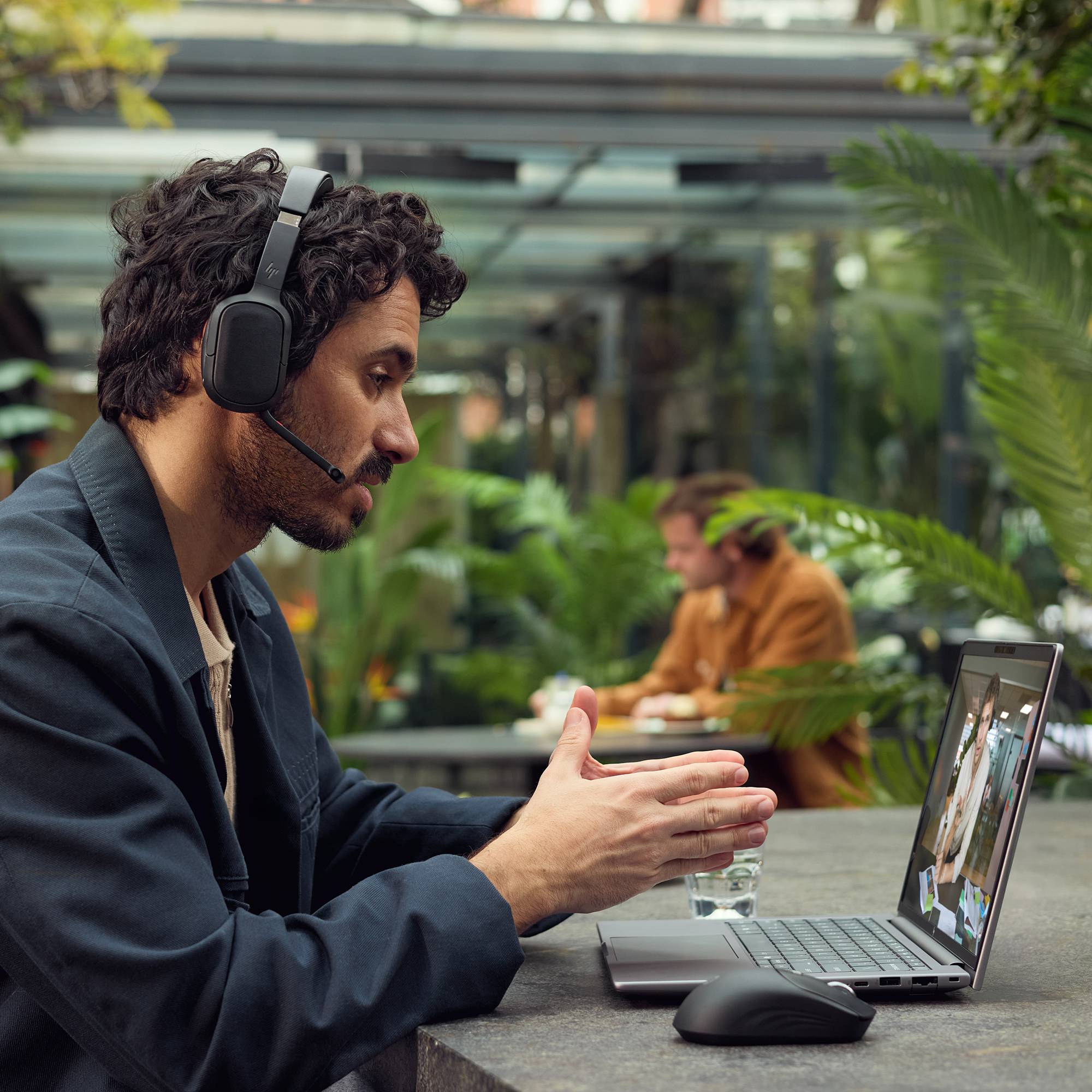 Ein Mann mit einem Kopfhörer gestikuliert während eines Videoanrufs auf einem Laptop an einem Outdoor-Café, mit einer Maus und einem Glas Wasser neben ihm.
