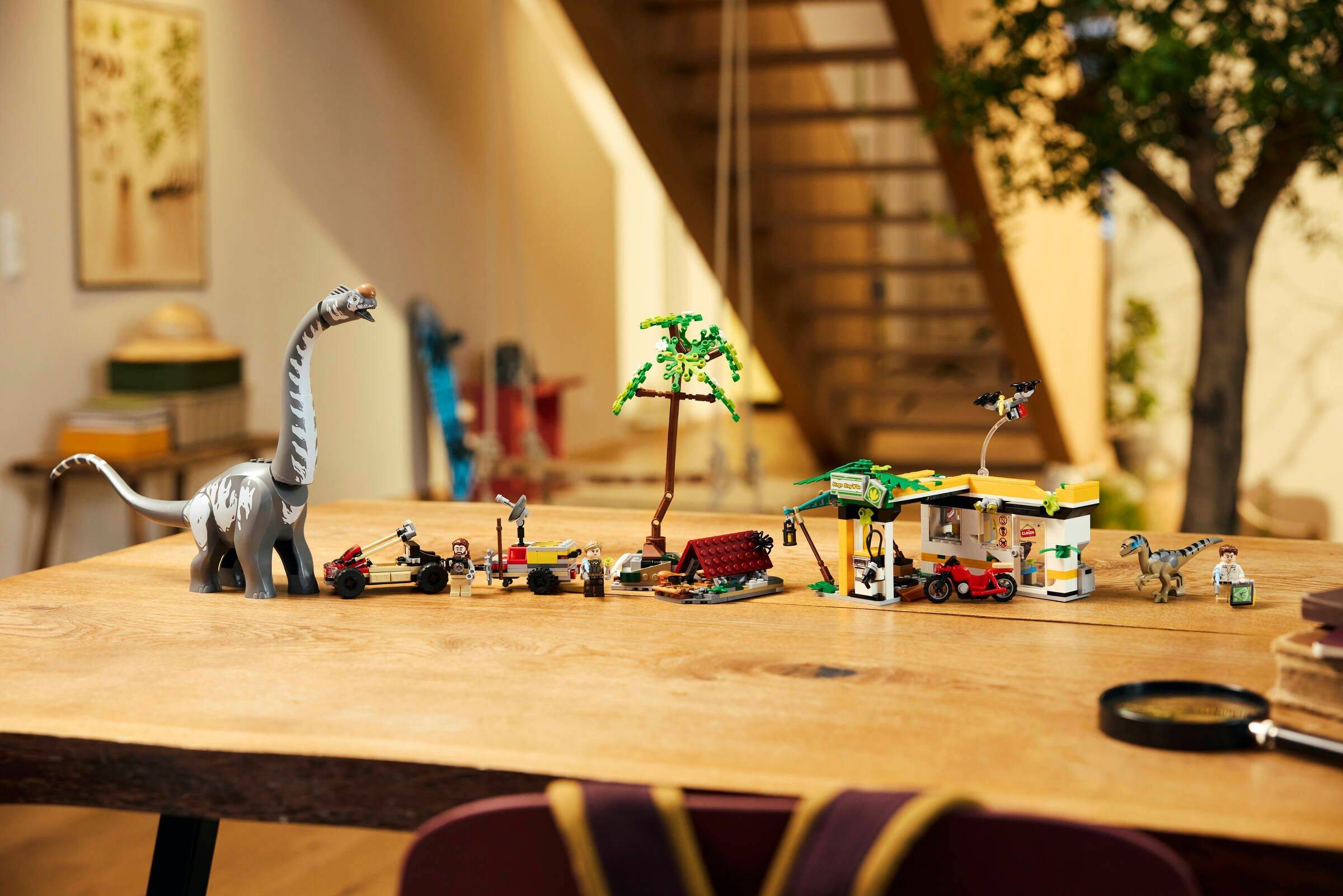 LEGO-Dinosaurier-Spielszene auf einem Holztisch, bestehend aus einem Langhalssaurier, kleinen Figuren, einer Palme und einer winzigen Station mit Fahrzeugen im Innenbereich.